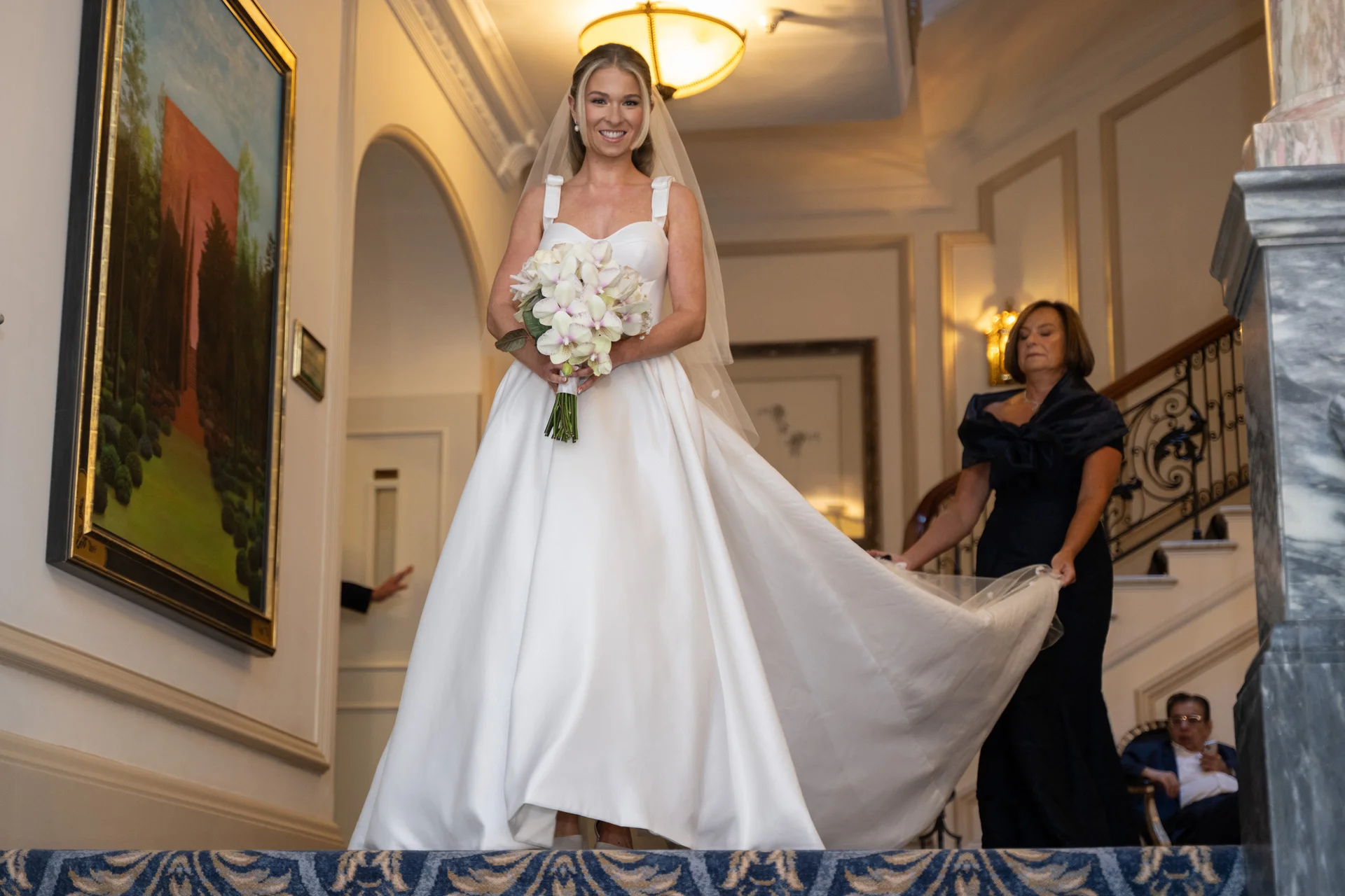 A radiant bride in a classic satin a line gown with thick straps smiles while holding a bouquet of white orchids as she descends a grand staircase, accompanied by her mother holding her dress train.