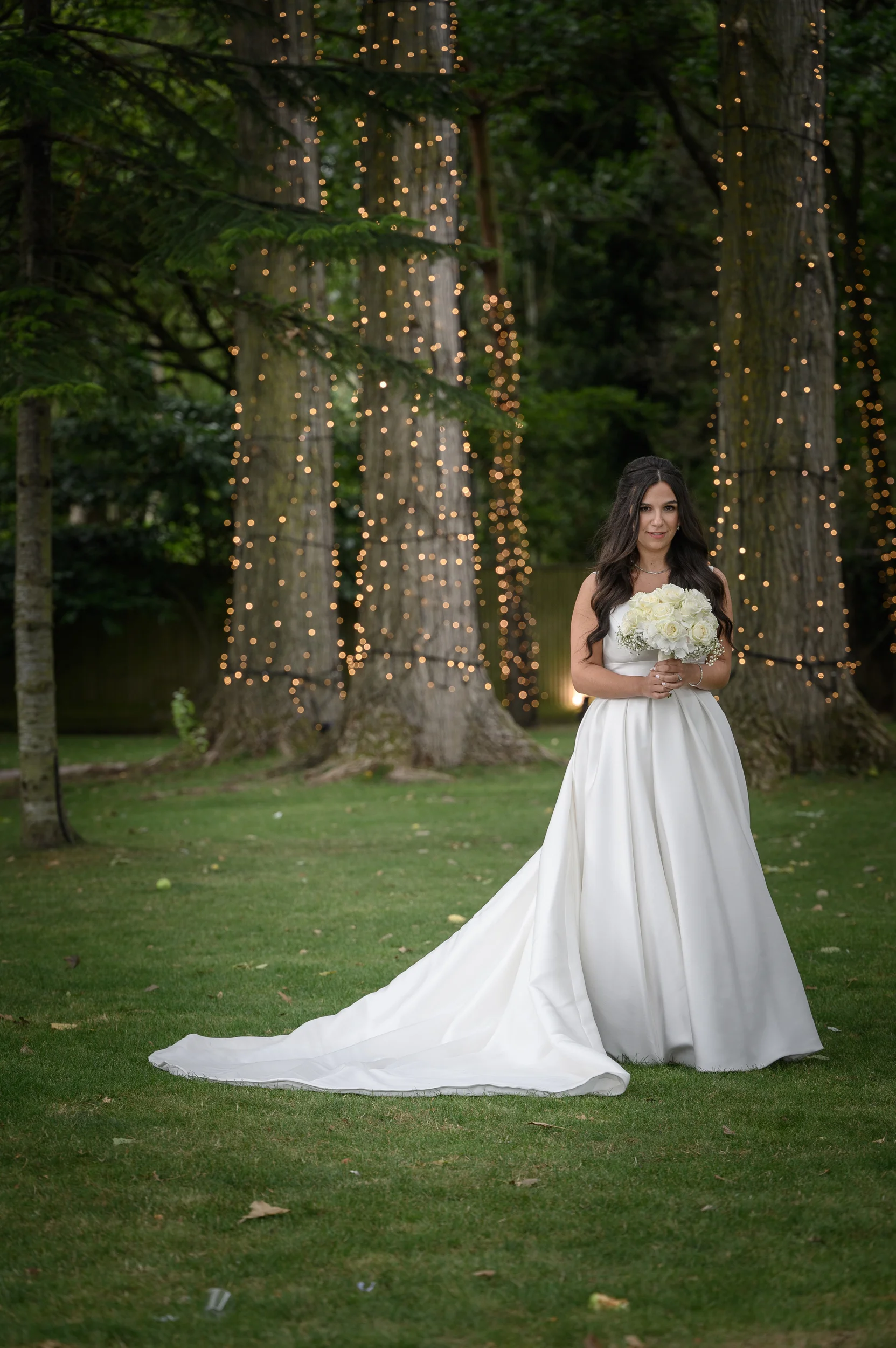 A glowing bride in a classic satin a-line gown with a long train poses gracefully in a woodland clearing illuminated by fairy lights on the trees.