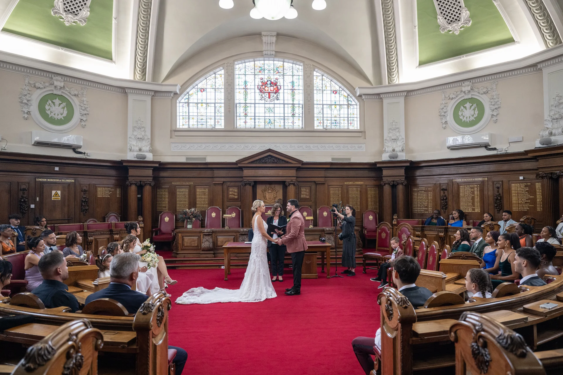 A bride and groom stand holding hands during their civil ceremony in a grand town hall with wood panelled walls, a red carpet, and stained glass windows.