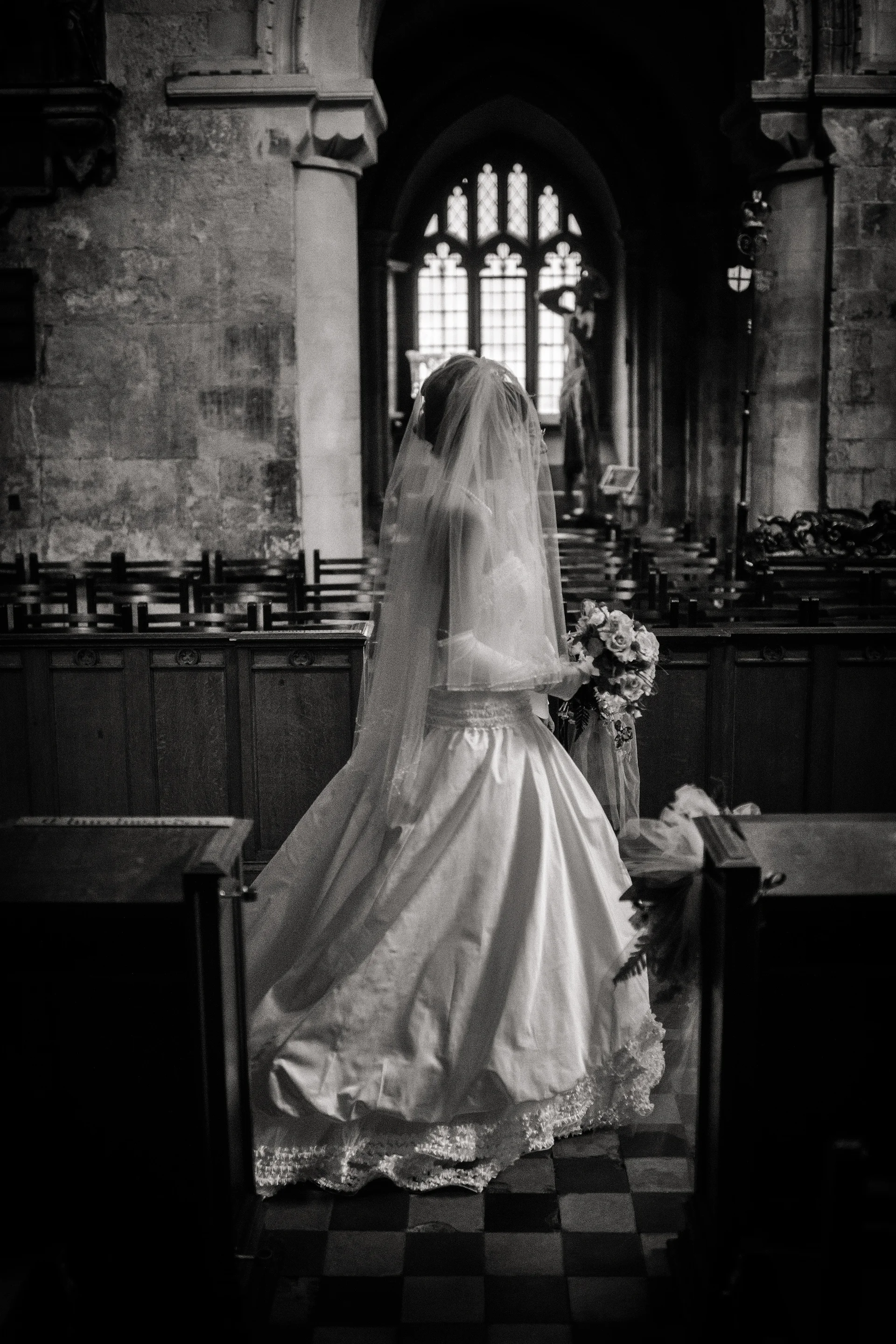 A bride in a classic white gown and veil walks down the aisle of an atmospheric stone church carrying a bouquet.