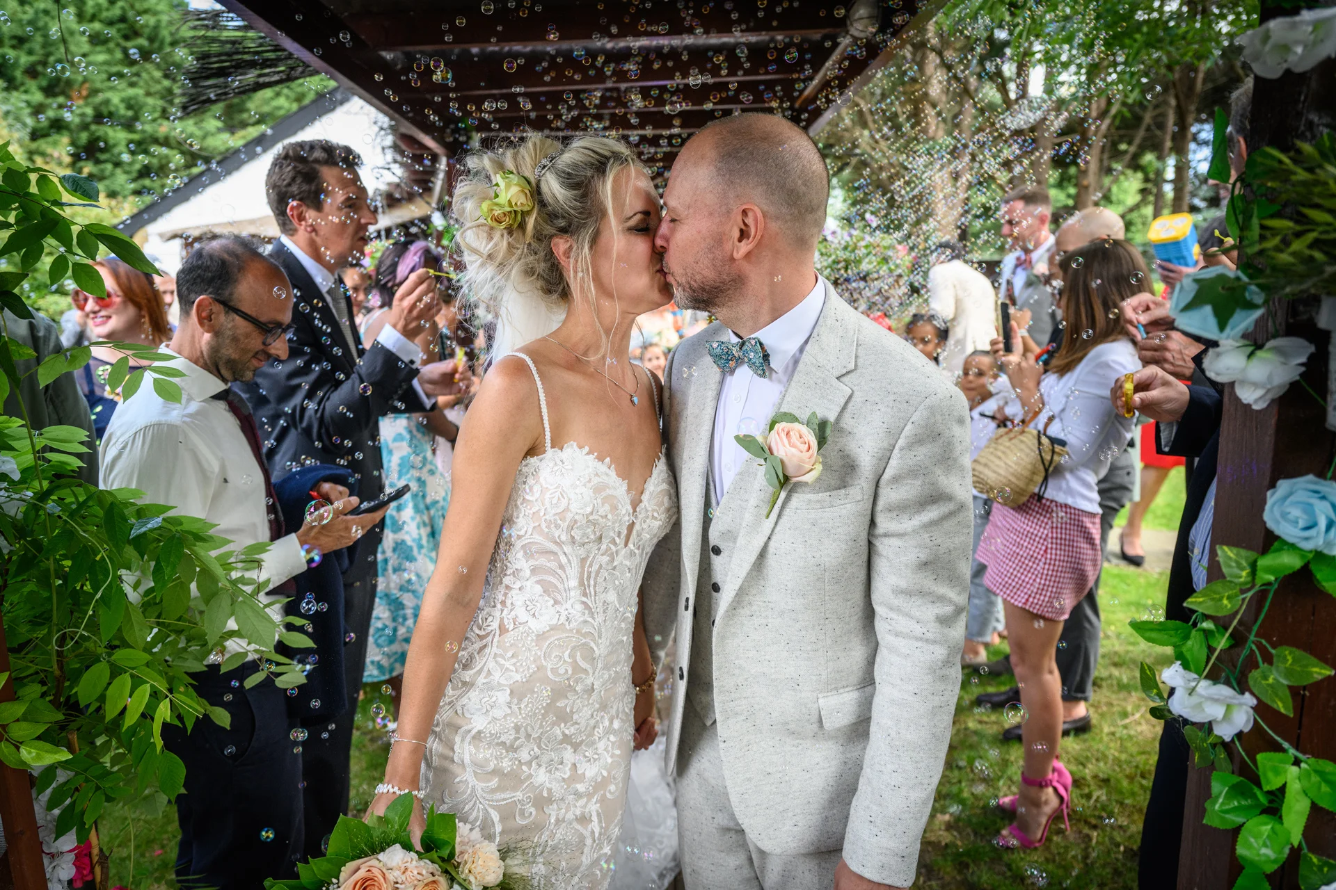Newlyweds share a romantic kiss beneath a wooden archway while bubbles float around them amidst cheering wedding guests.