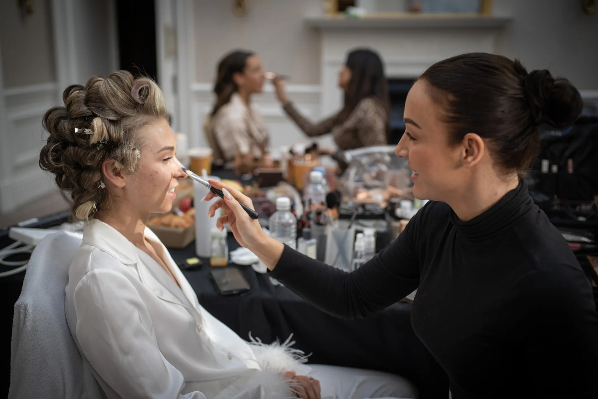 A bride with hair rollers sits in a white robe while a makeup artist carefully applies foundation to her nose during morning bridal preparations.