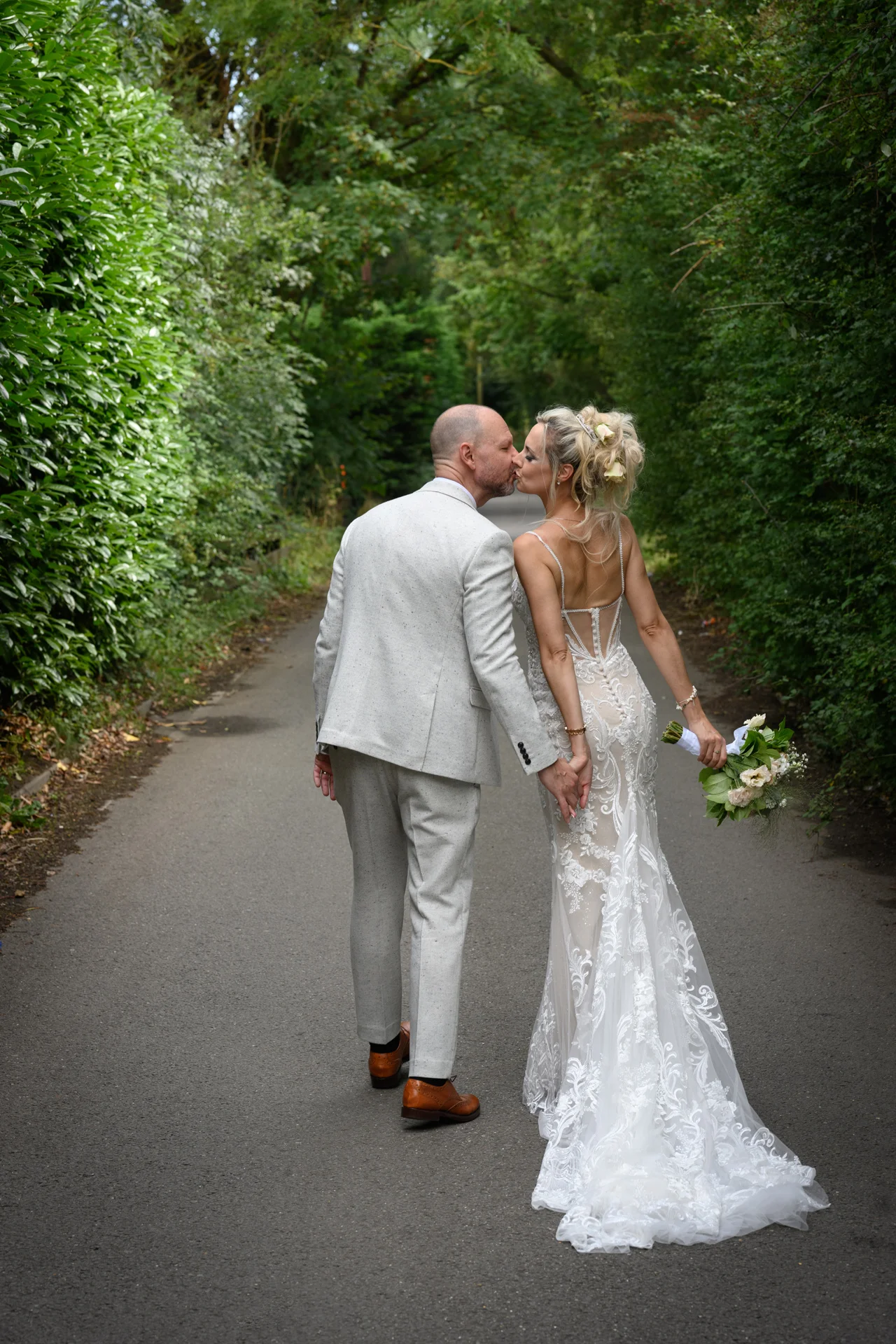 Newlyweds share a tender kiss while walking hand in hand down a tree-lined lane on their wedding day.