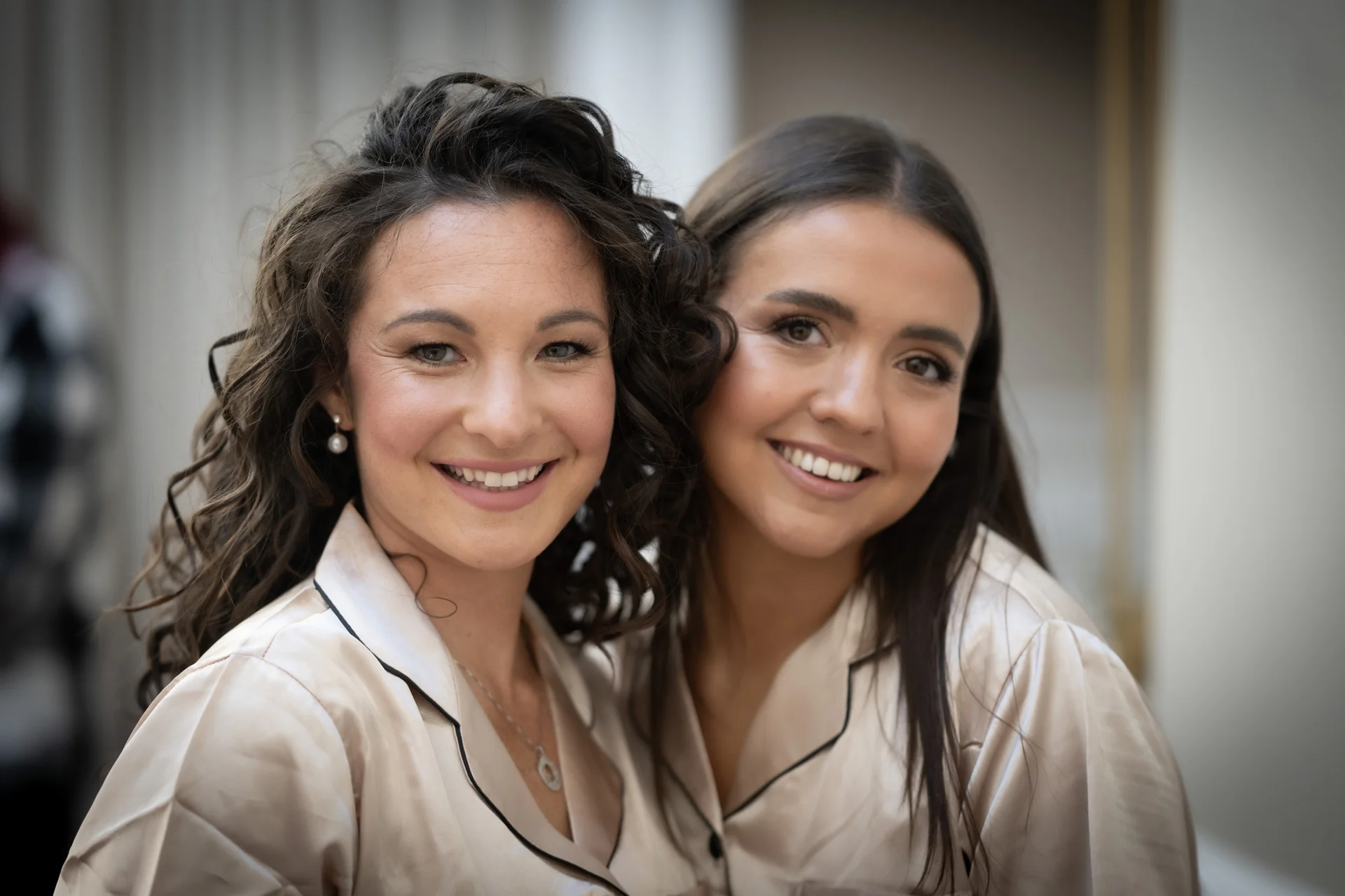 Two smiling women wearing matching satin pyjamas and pearl earrings pose closely together during morning bridal preparations.