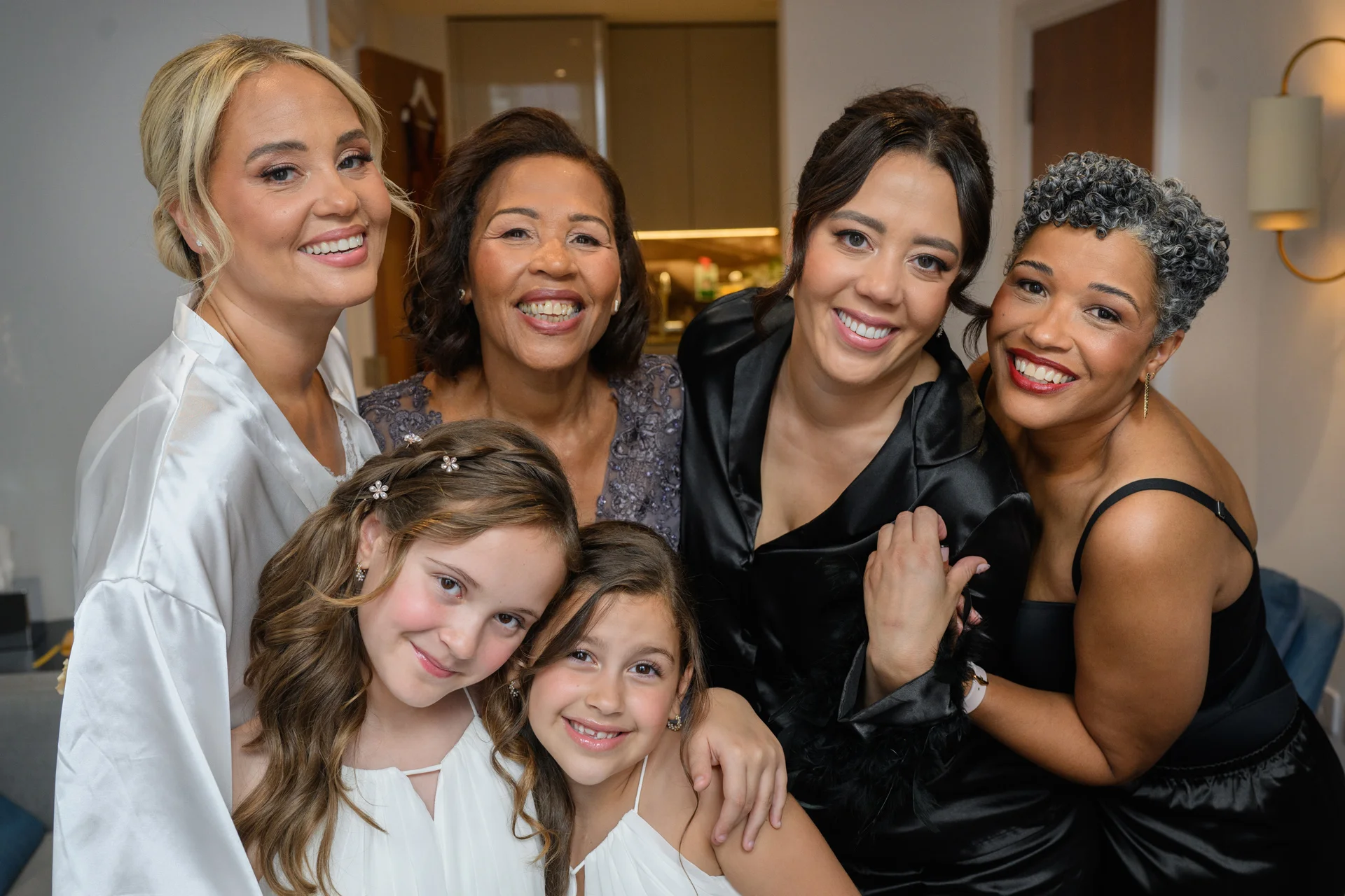 A multi-generational group of smiling women and young flower girls pose together indoors while getting ready for the wedding celebration.
