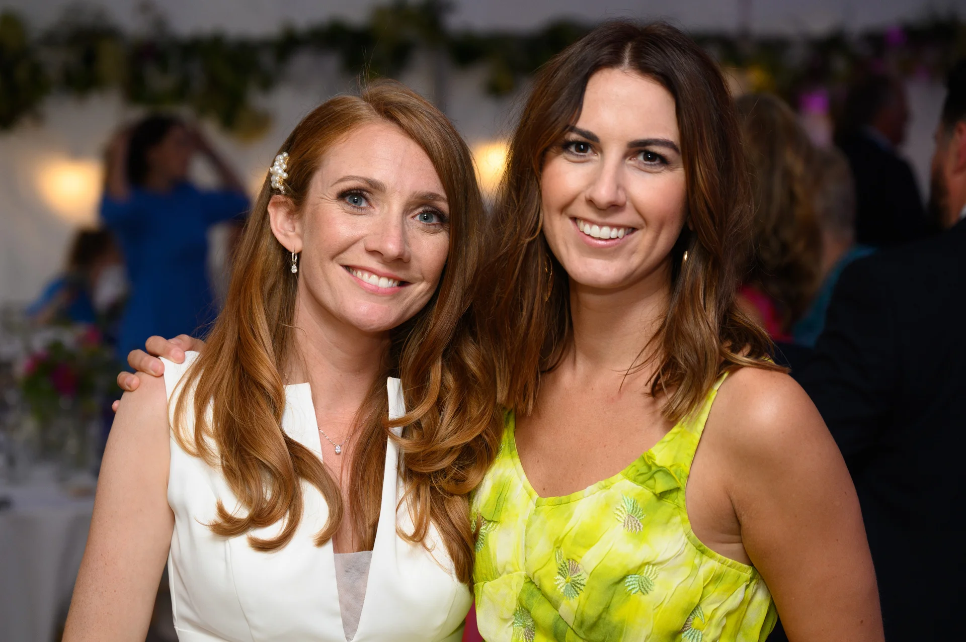 Two smiling female wedding guests pose together happily at an evening reception with soft ambient lighting in the background.