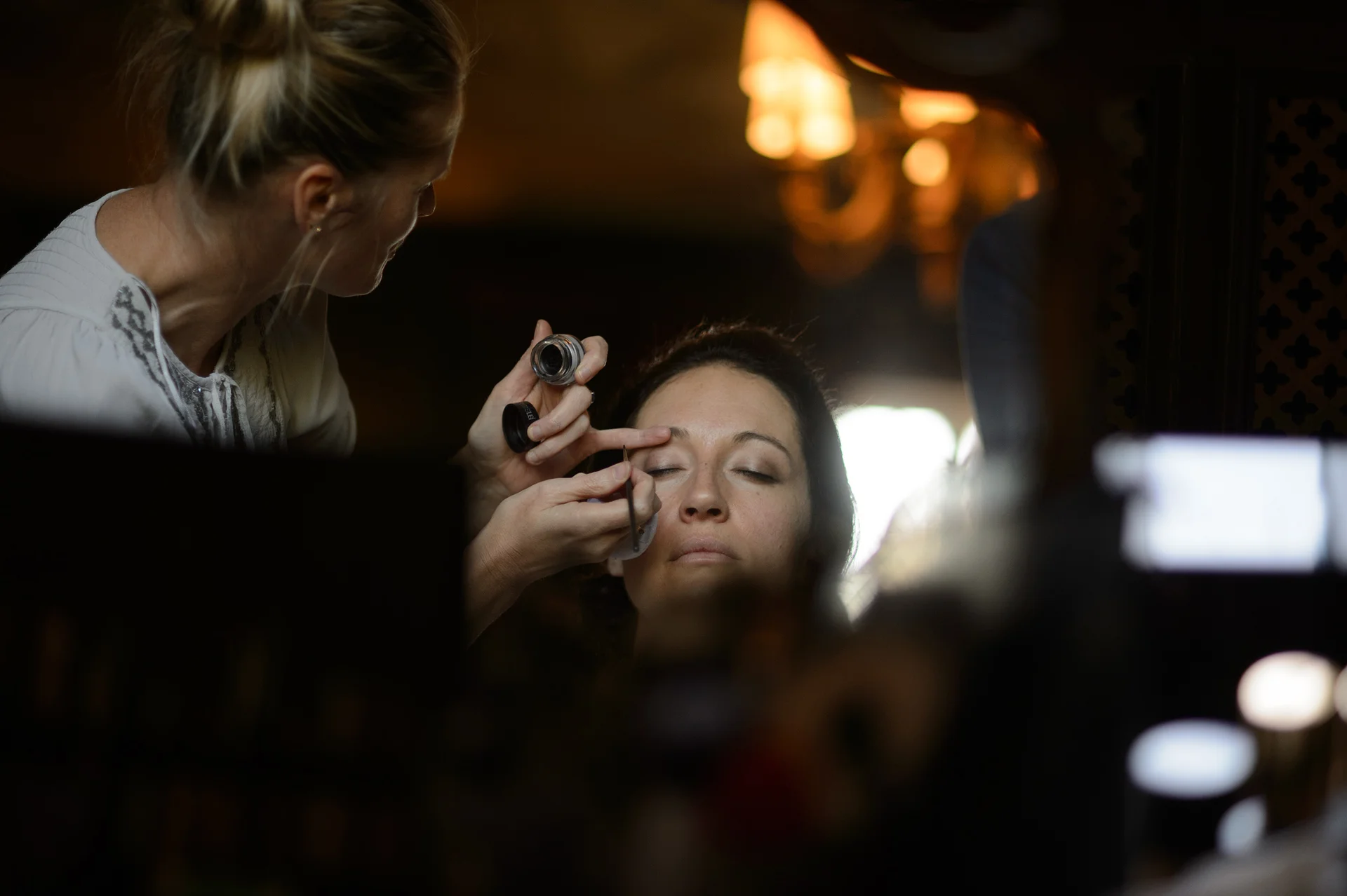 A makeup artist delicately applies eyeliner to a bride during morning preparations in a warmly lit room.