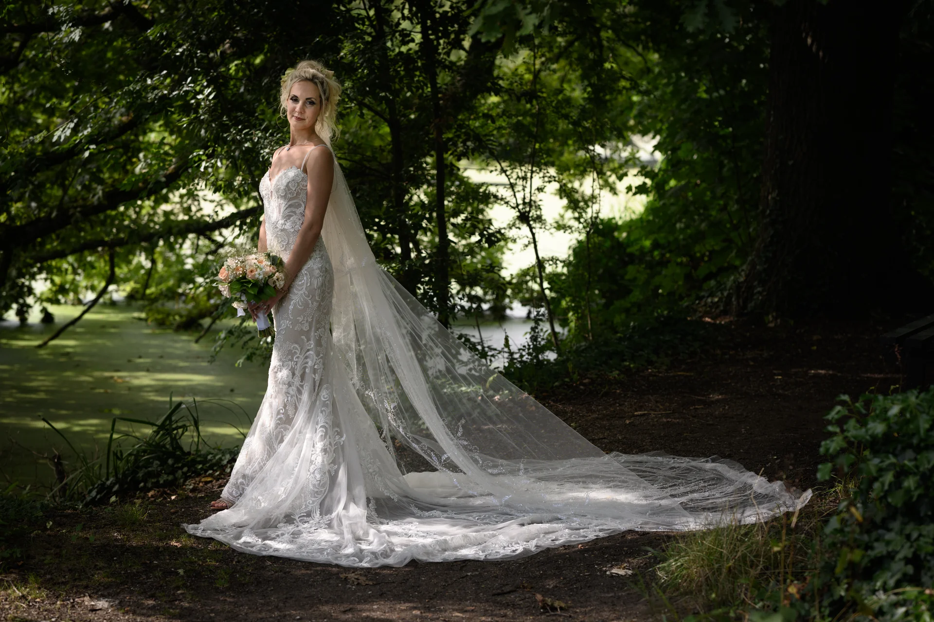 A stunning bride in a lace strapless wedding gown and cathedral veil poses gracefully in a sunlit woodland setting, holding a delicate bouquet.