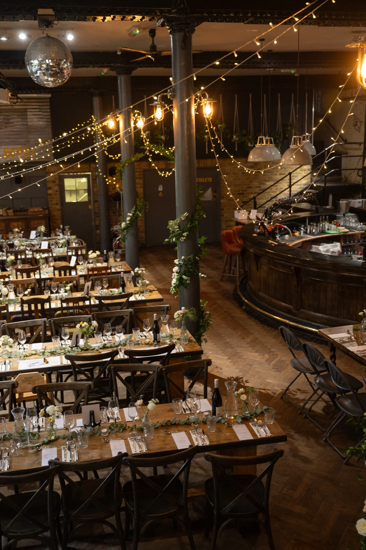 An overhead view of a warmly lit industrial wedding venue featuring long wooden banquet tables adorned with greenery runners, bud vases, and taper candles beneath a canopy of fairy lights and a hanging disco ball.