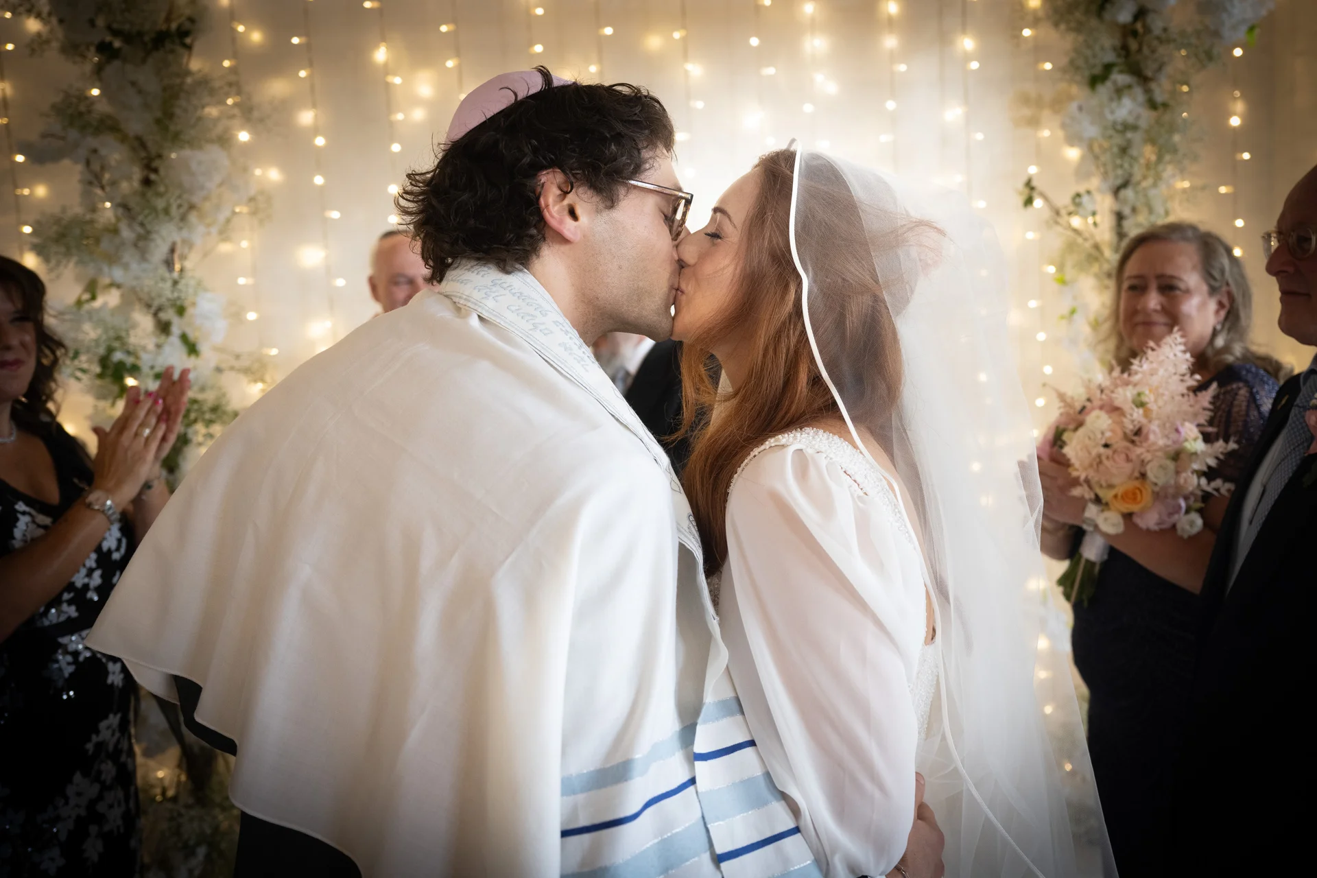A newly married couple share their first kiss during a Jewish wedding ceremony, framed by a glowing fairy light backdrop and smiling guests.