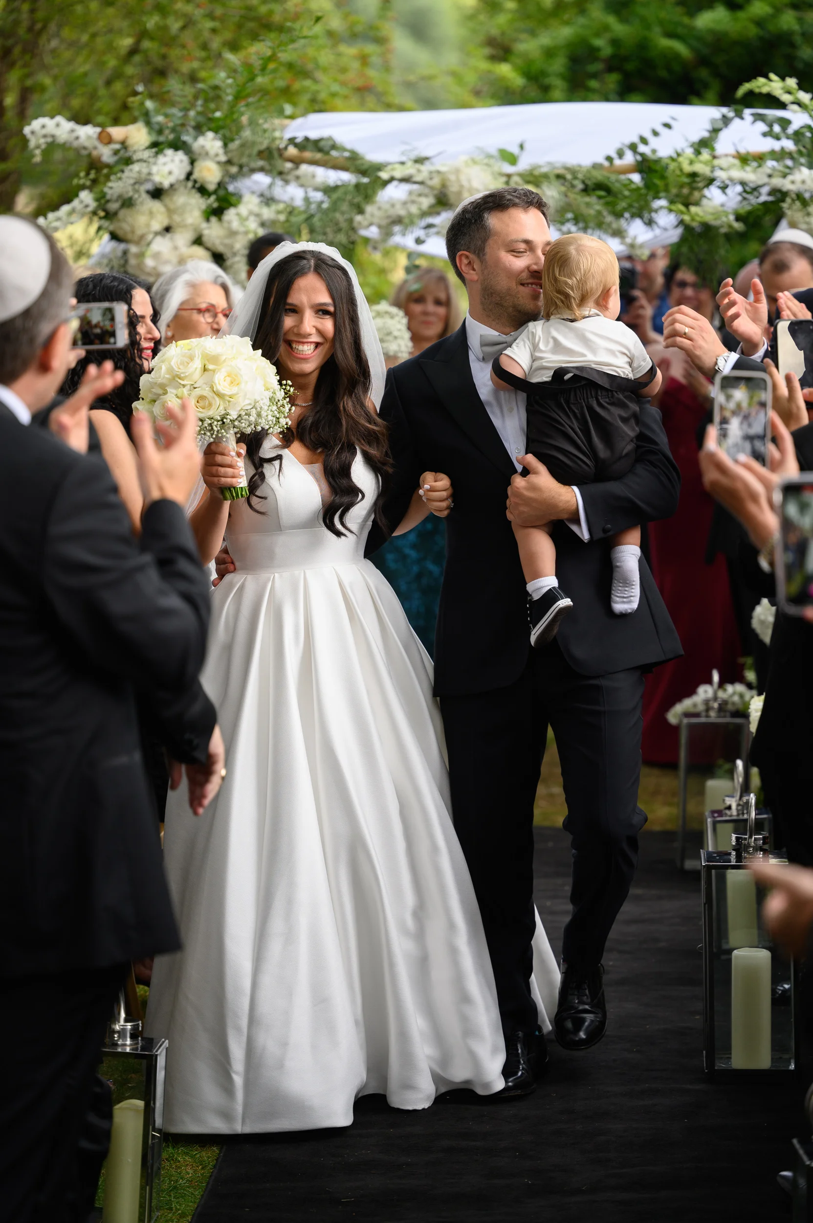 A joyful newlywed couple walks down the aisle hand in hand after their outdoor Jewish wedding ceremony, with the groom carrying a young child while guests applaud and film on their phones.