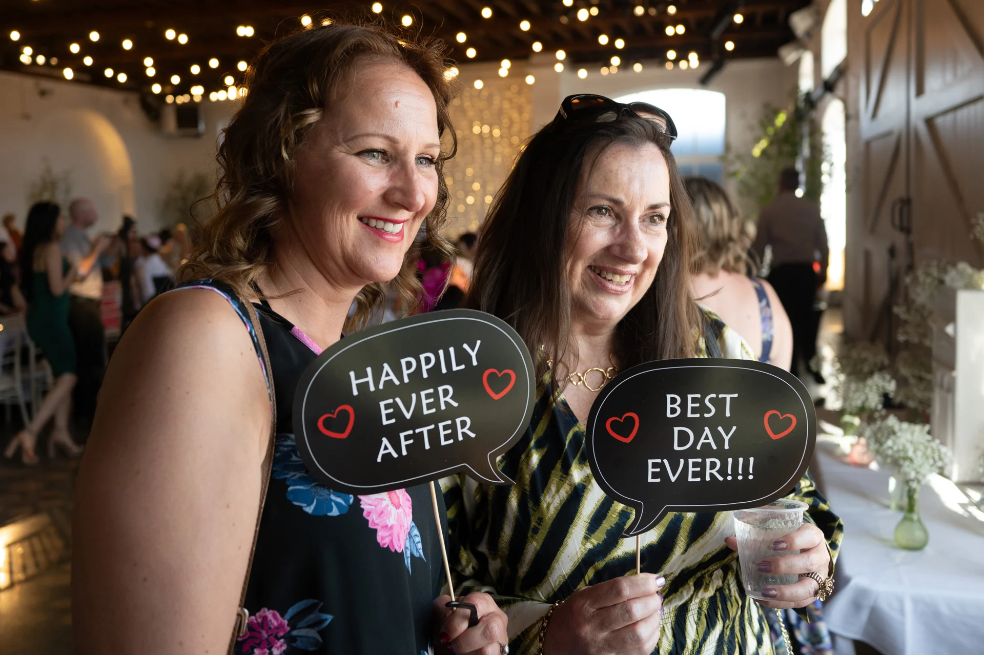 Two smiling female wedding guests hold speech bubble photo booth props reading happily ever after and best day ever inside a rustic barn venue.