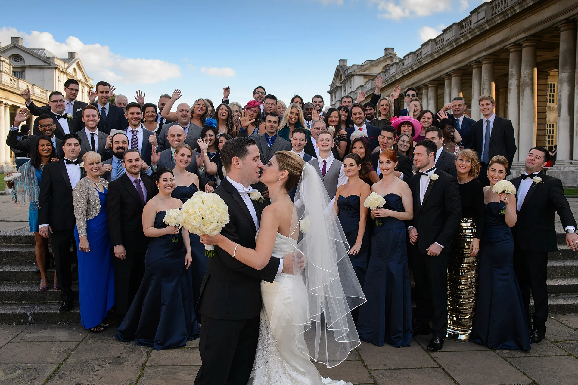 The bride and groom share a kiss in the foreground while their large wedding party and guests celebrate and wave on the grand stone steps of a stately home.