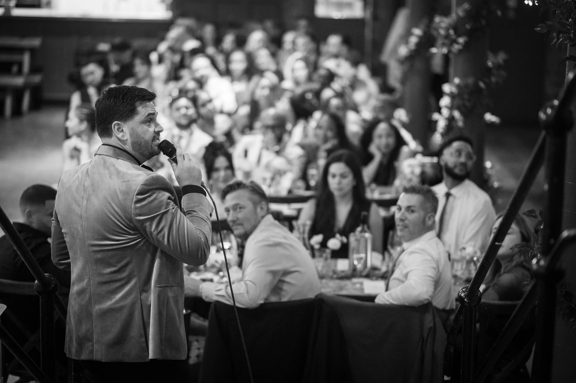 A groom delivers a speech into a microphone during the wedding reception while seated guests look on and smile in black and white.