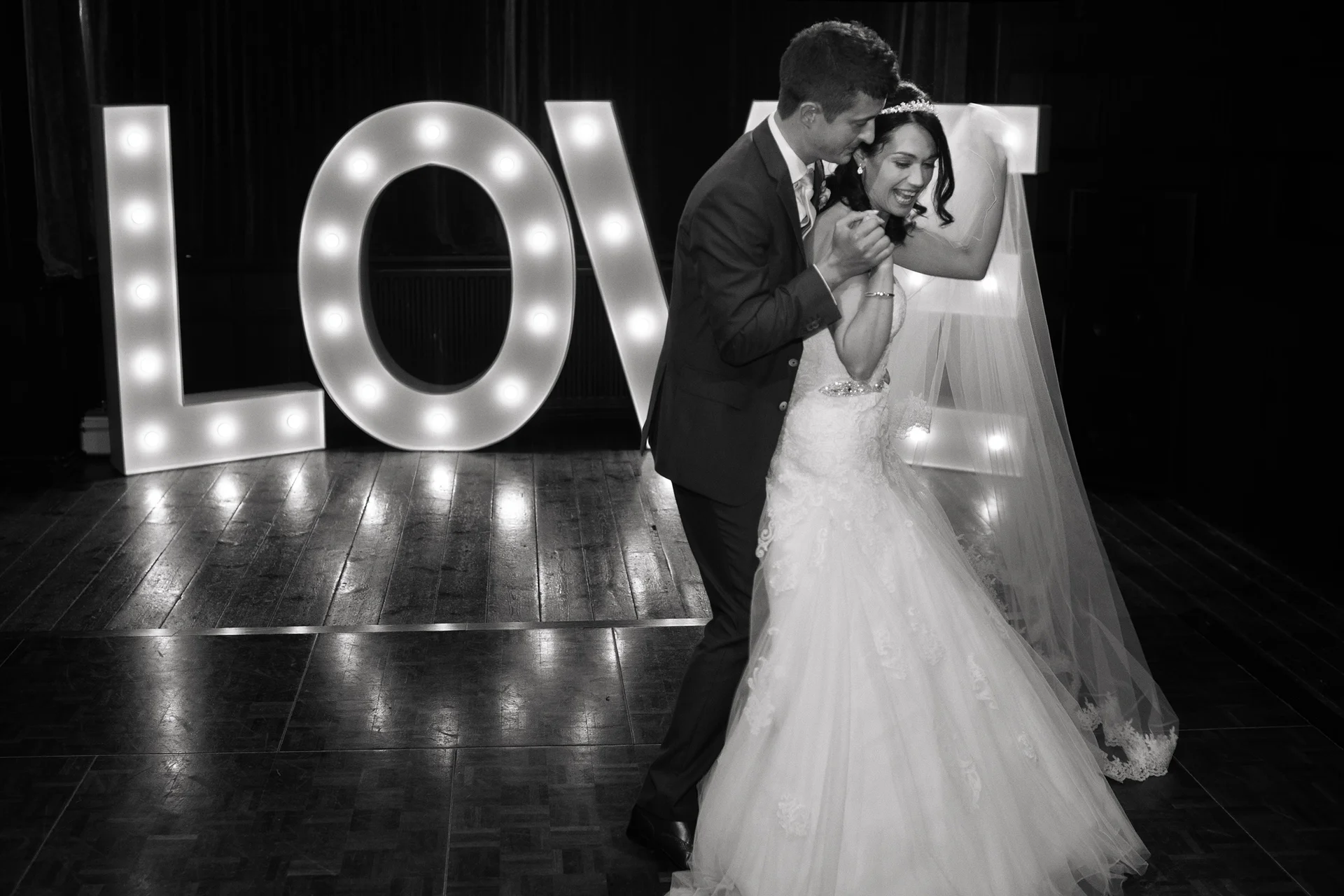 A newlywed couple shares a joyful first dance on a polished wooden dance floor in front of large illuminated letters spelling LOVE.
