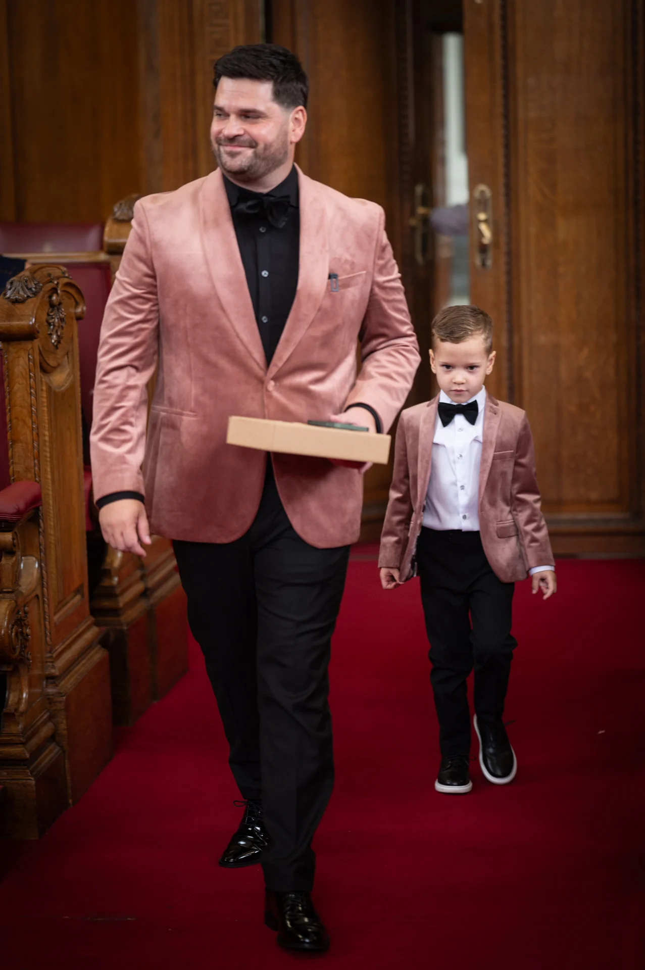 A groom in a pink velvet blazer and black shirt walks down a red-carpeted aisle alongside a young page boy wearing matching attire.