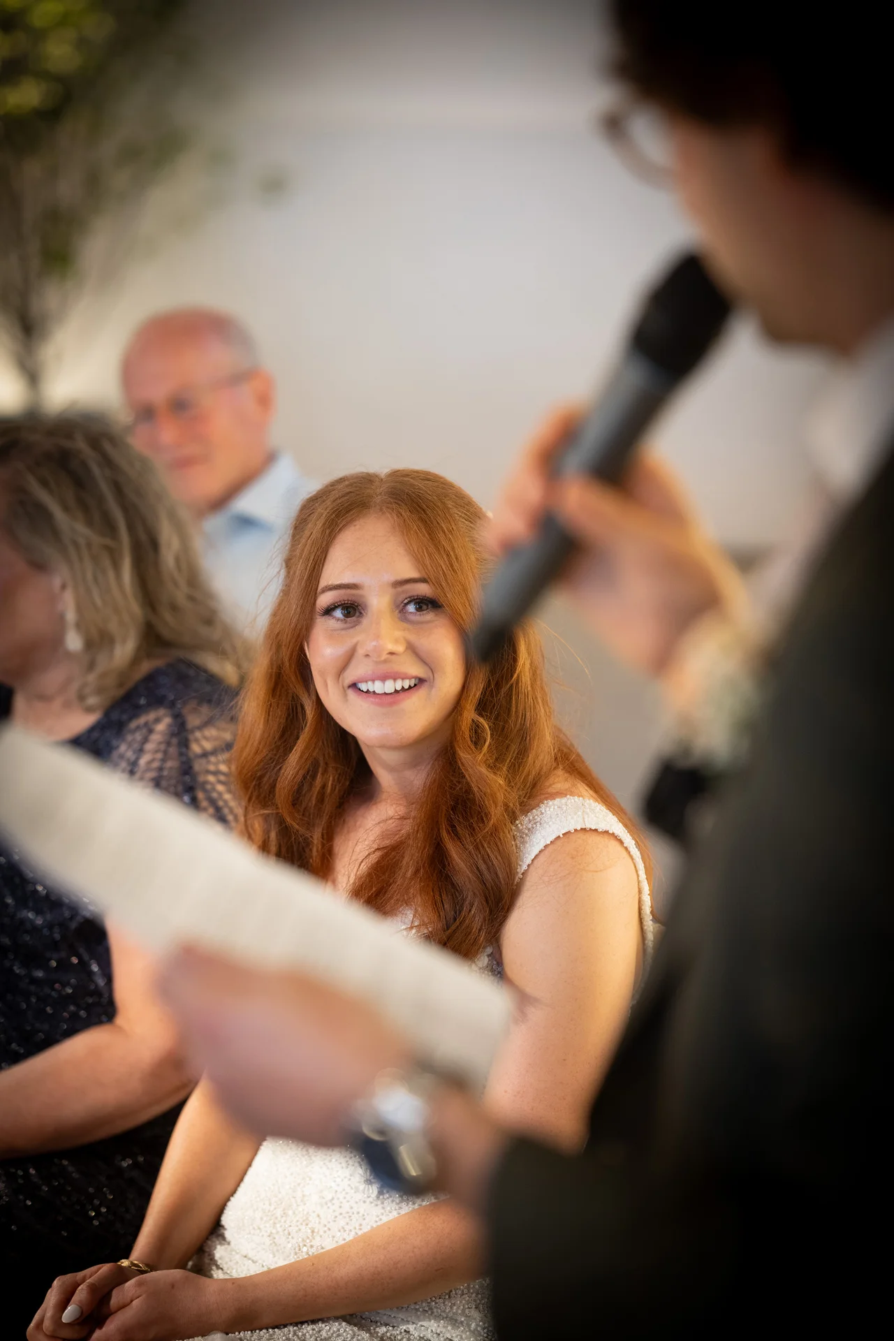 A smiling red-haired bride with half-up wavy hair looks towards a speaker holding a microphone during the wedding speeches.