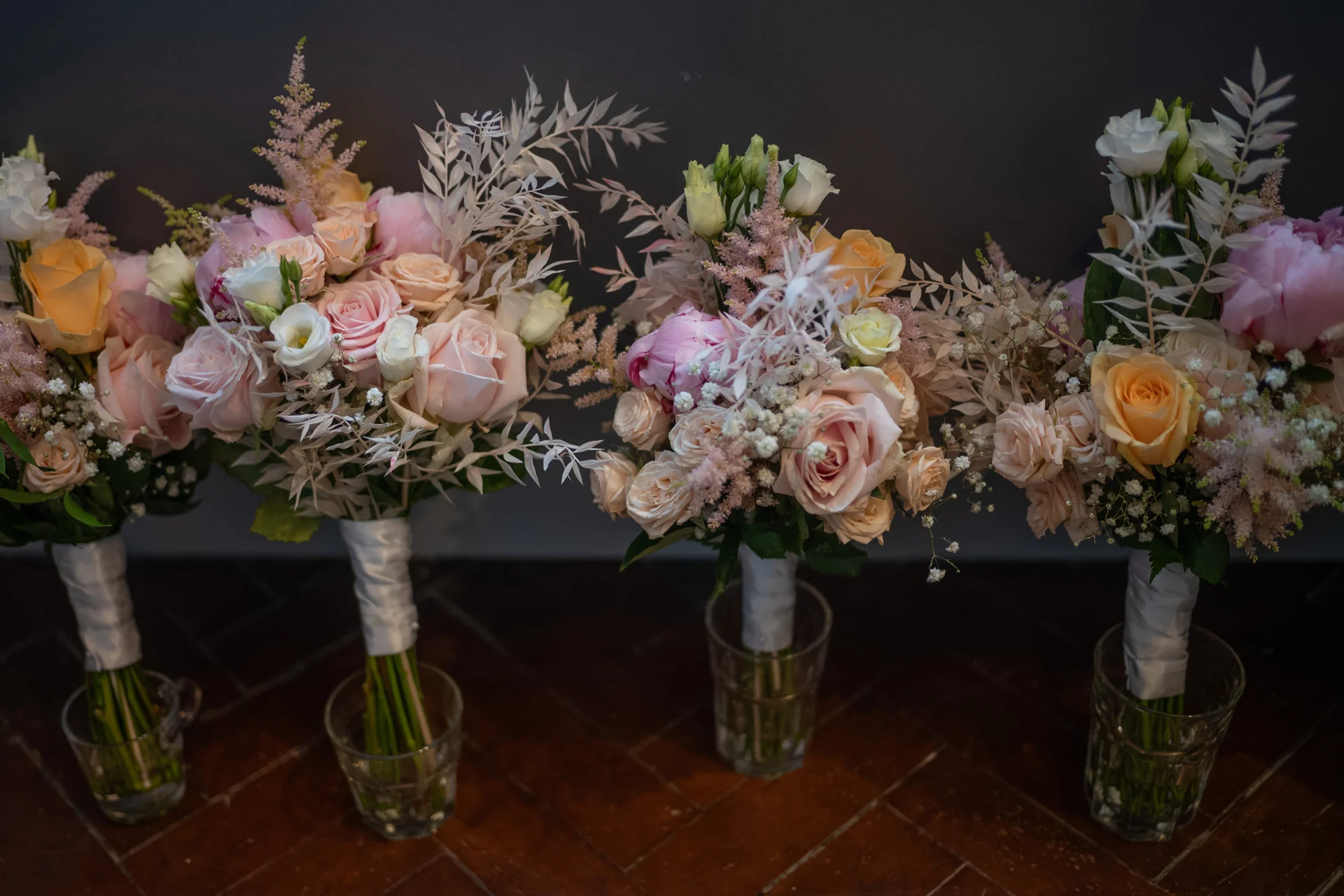 Three stunning hand-tied bridal bouquets featuring soft pink and peach roses, delicate peonies, and bleached fern foliage displayed in glass tumblers on a wooden floor.