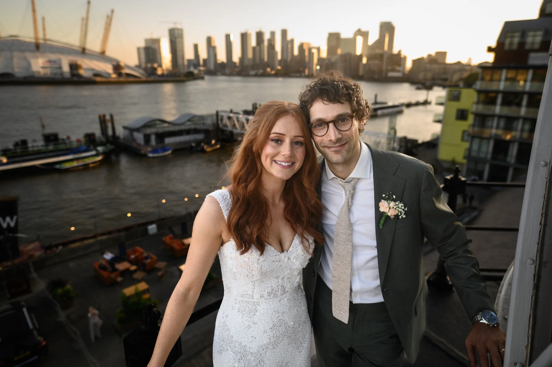 A smiling bride and groom stand on a rooftop overlooking the London skyline and river at sunset during their wedding celebration.
