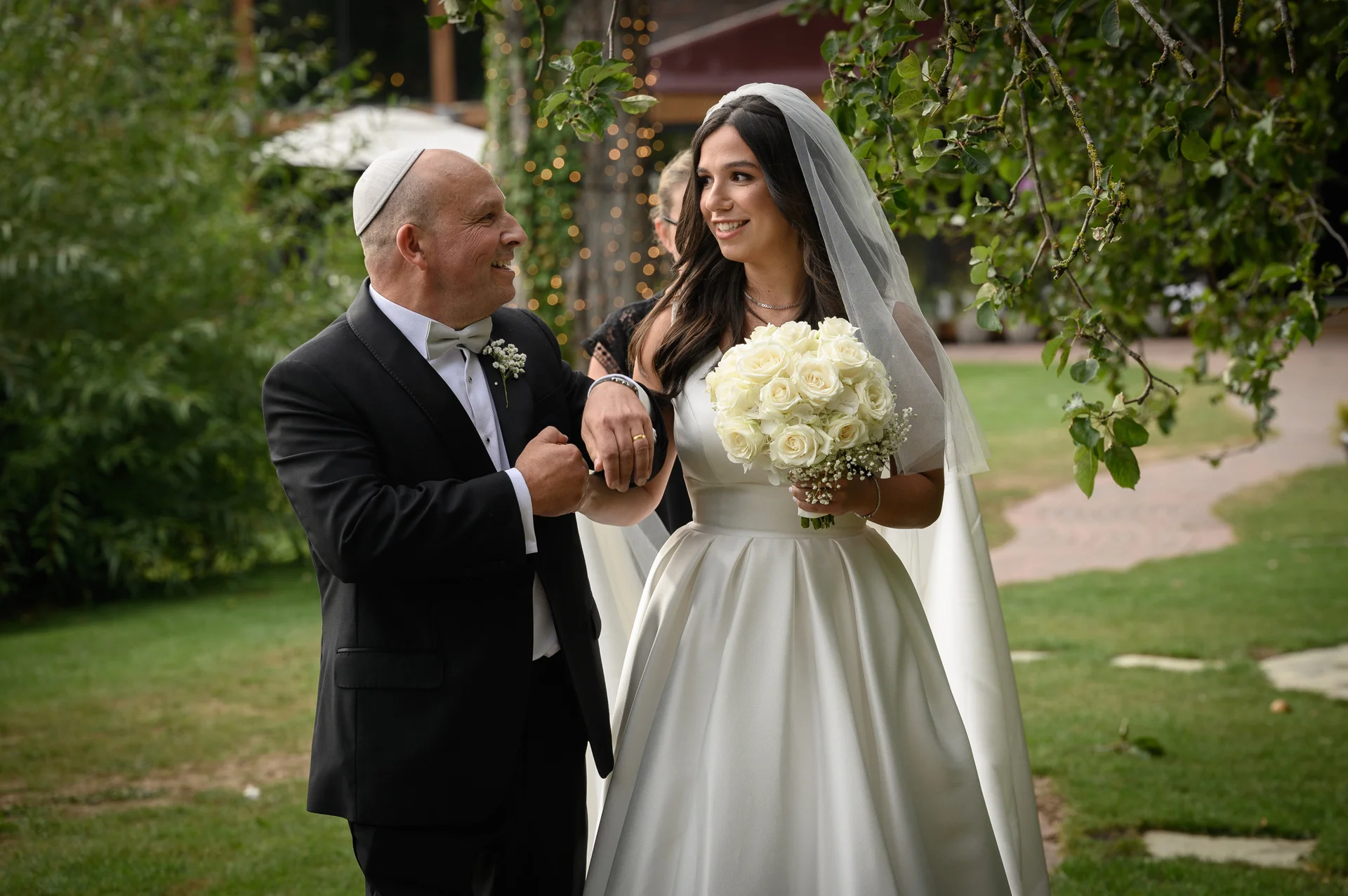 A smiling bride holding a bouquet of white roses walks arm-in-arm with her father outdoors during a wedding ceremony.