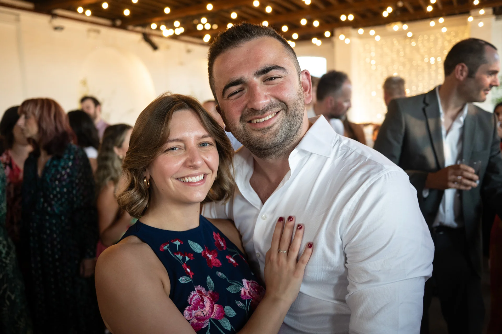 A happy couple smiling together at a wedding reception under a ceiling draped with twinkling string lights.