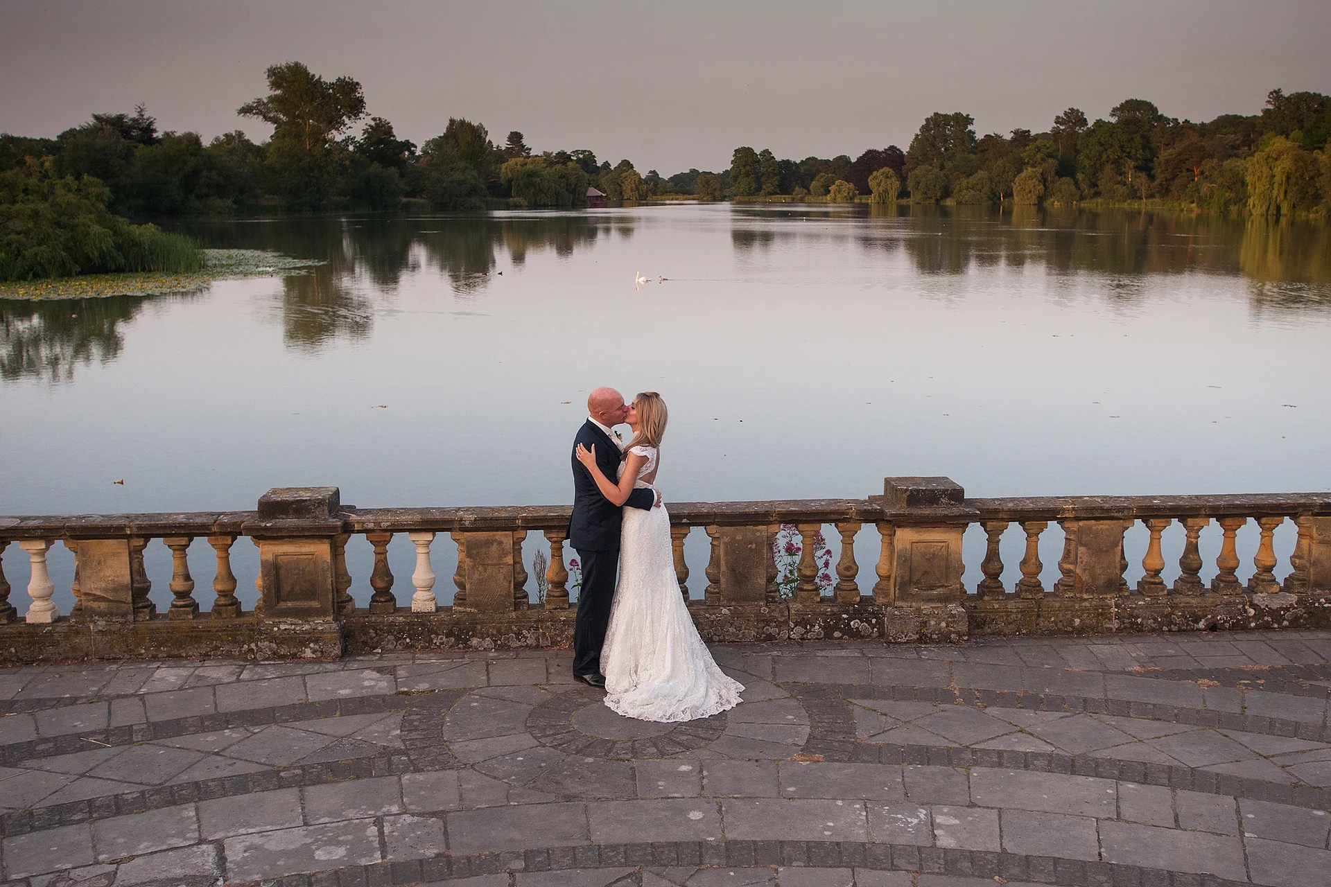 A newlywed couple share a romantic kiss by an ornamental lake on a stone terrace at dusk.