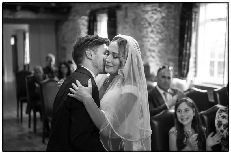 A groom gently kisses the bride on the cheek immediately after their ceremony inside a rustic stone venue, with guests smiling and applauding in the background.