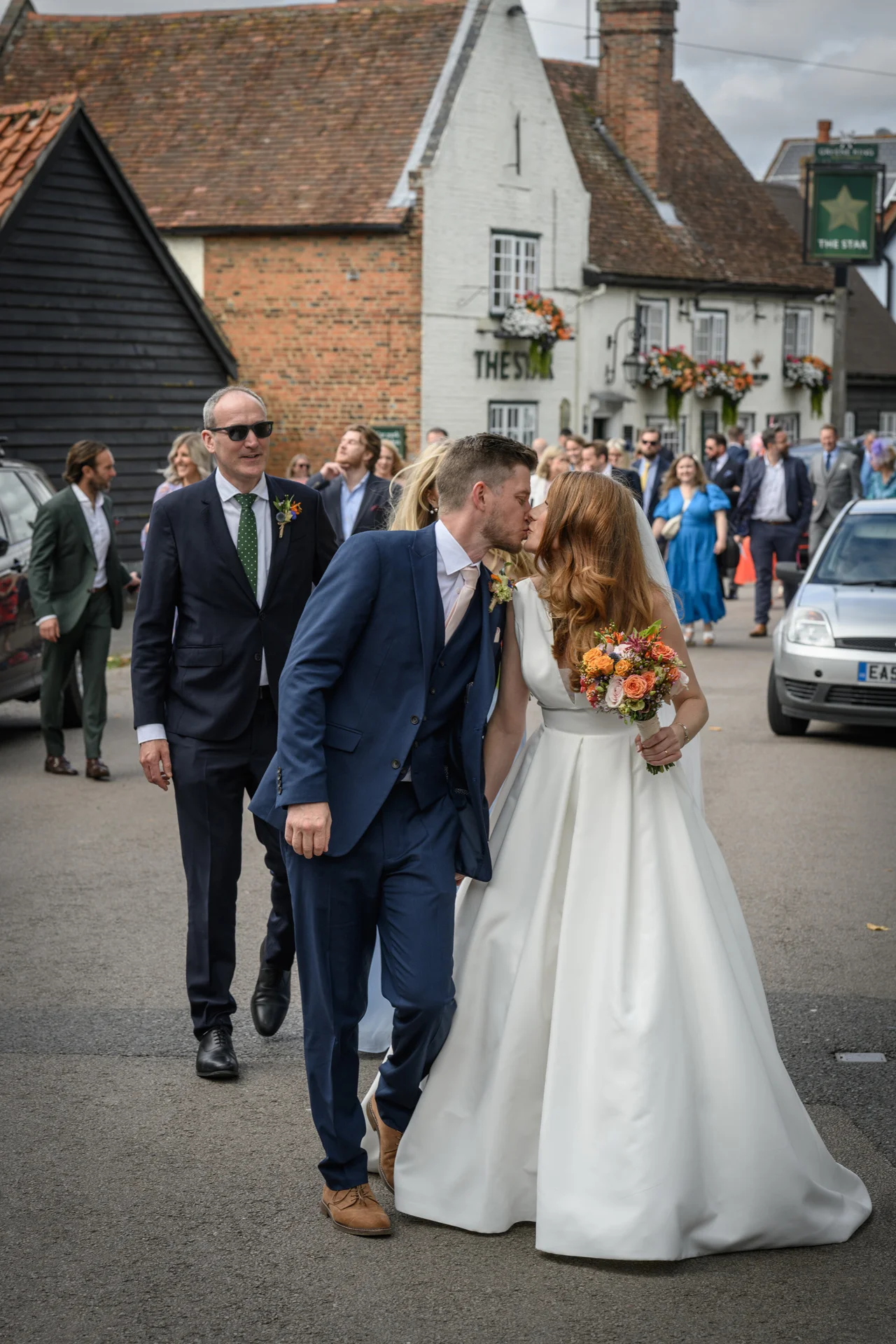 A newlywed couple shares a kiss while walking down a village street outside a pub, followed by wedding guests and family members.