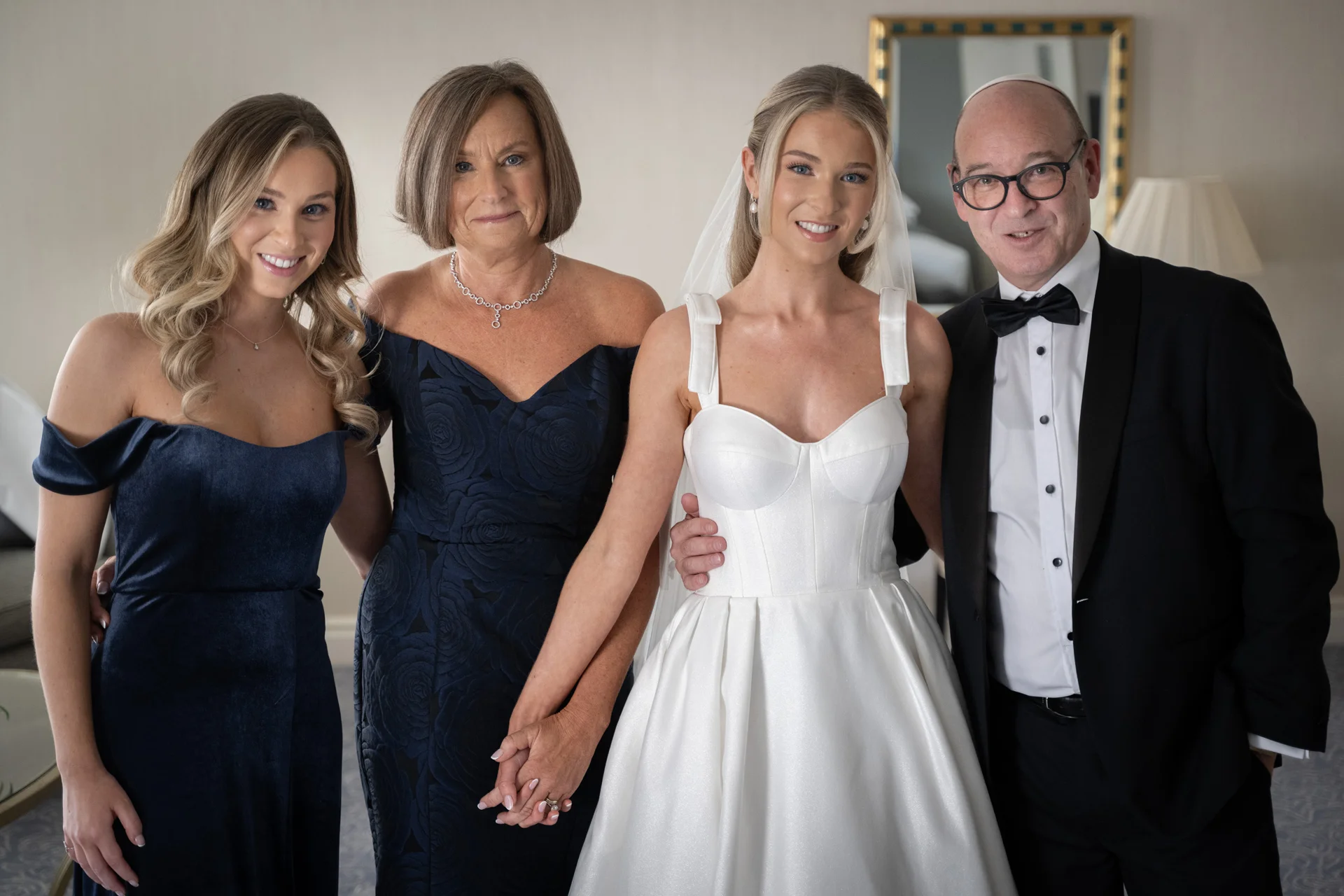 A smiling bride in a structured white gown stands with her mother, father in a tuxedo, and another relative, holding hands indoors.