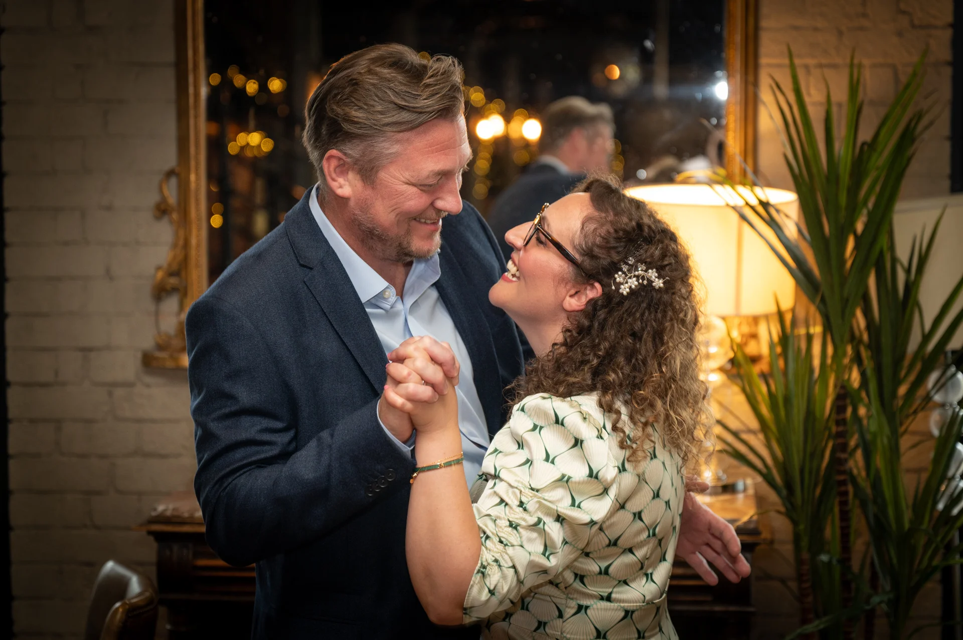 An older couple lovingly dancing and laughing together indoors at night, illuminated by warm ambient lighting near a mirror and exposed brick wall.