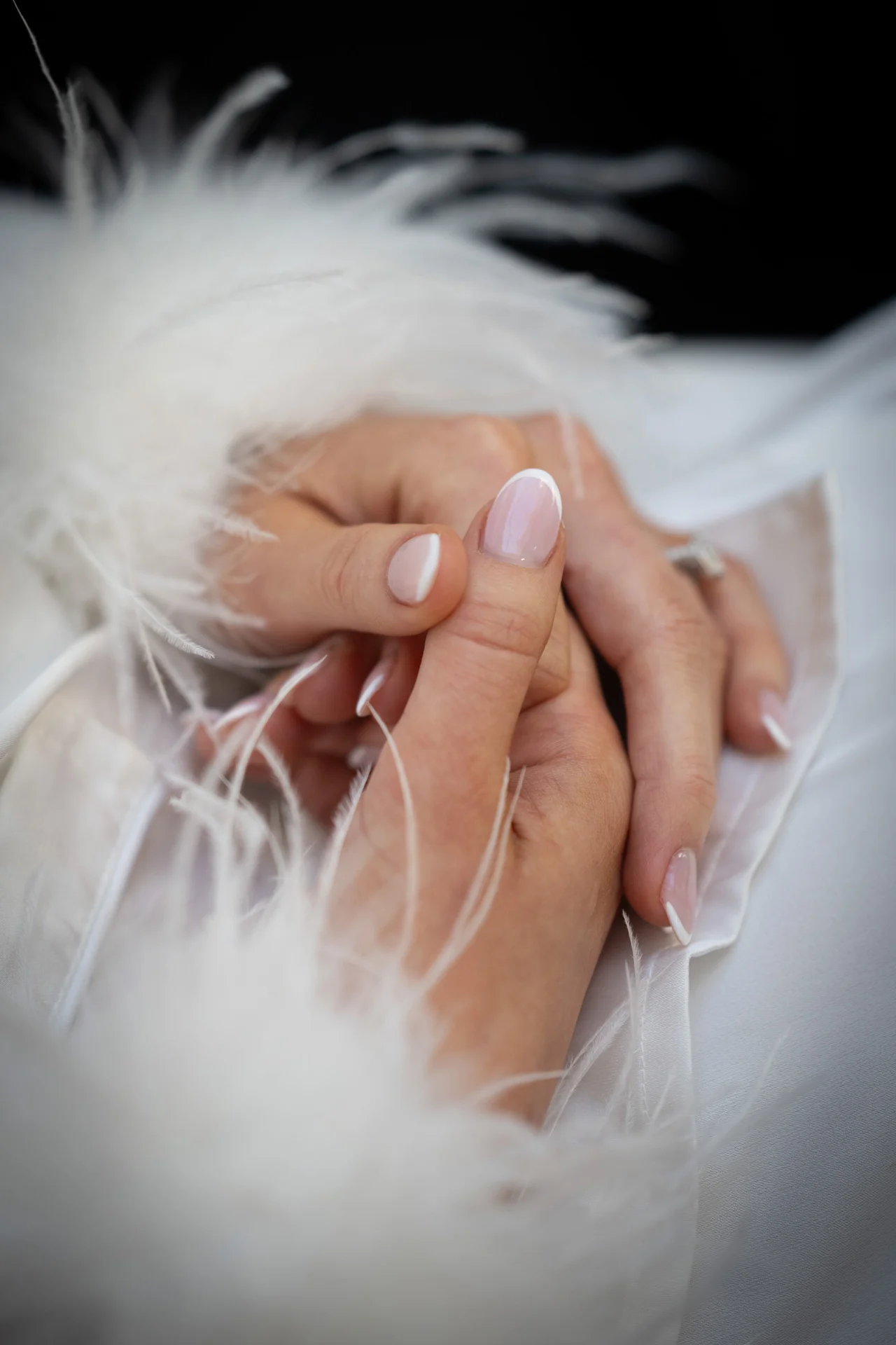 An intimate close-up shot captures hands holding gently, adorned with a classic french manicure and delicate feather-trimmed cuffs.