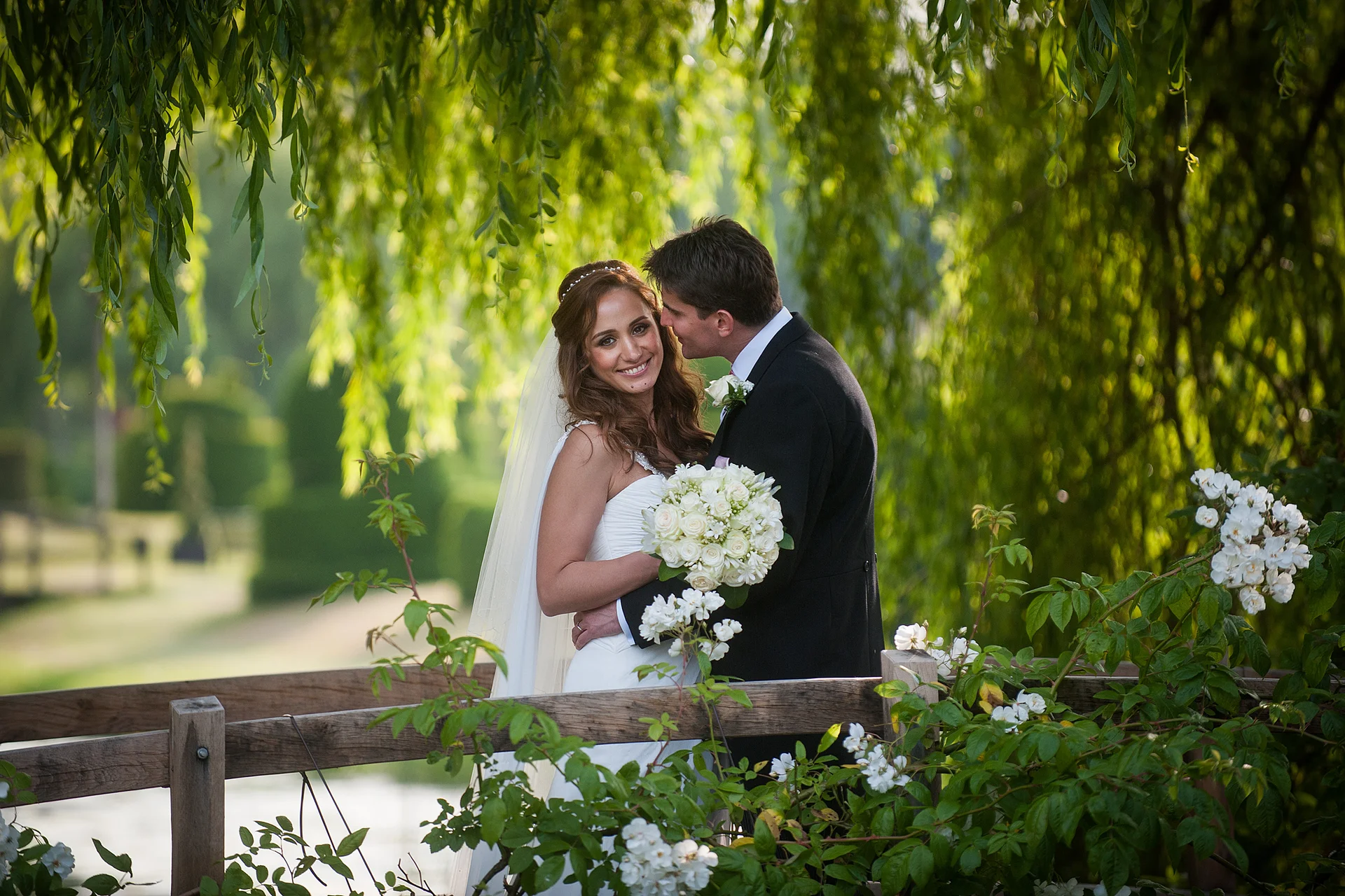 A smiling bride and groom embrace affectionately on a wooden bridge, framed by weeping willow branches and glowing natural light.