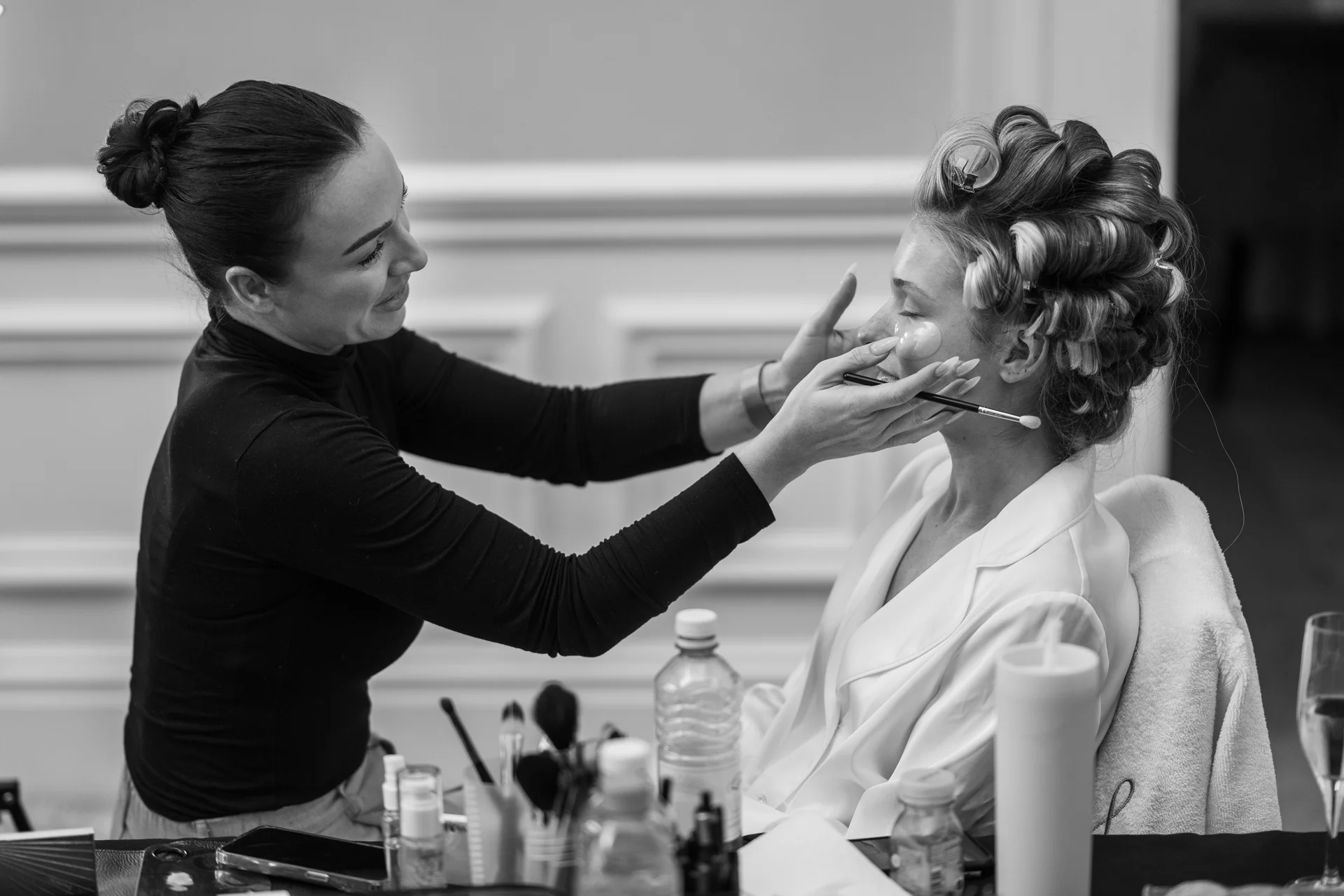 A professional makeup artist applies cosmetics to a bride who has hair rollers in her hair during bridal preparations.