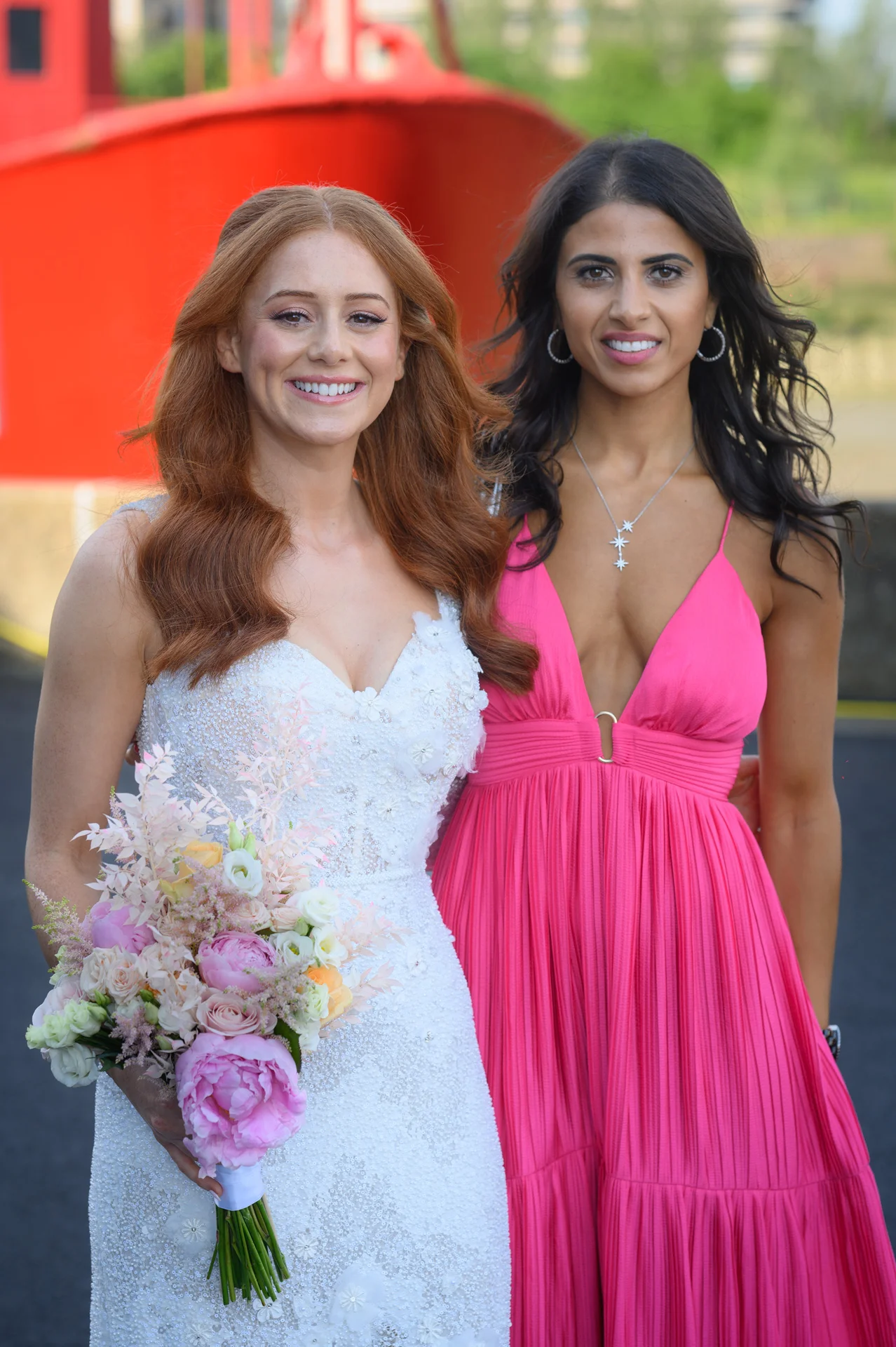A smiling red-haired bride holding a pastel peony and rose bouquet poses next to a bridesmaid wearing a vibrant pink pleated gown.