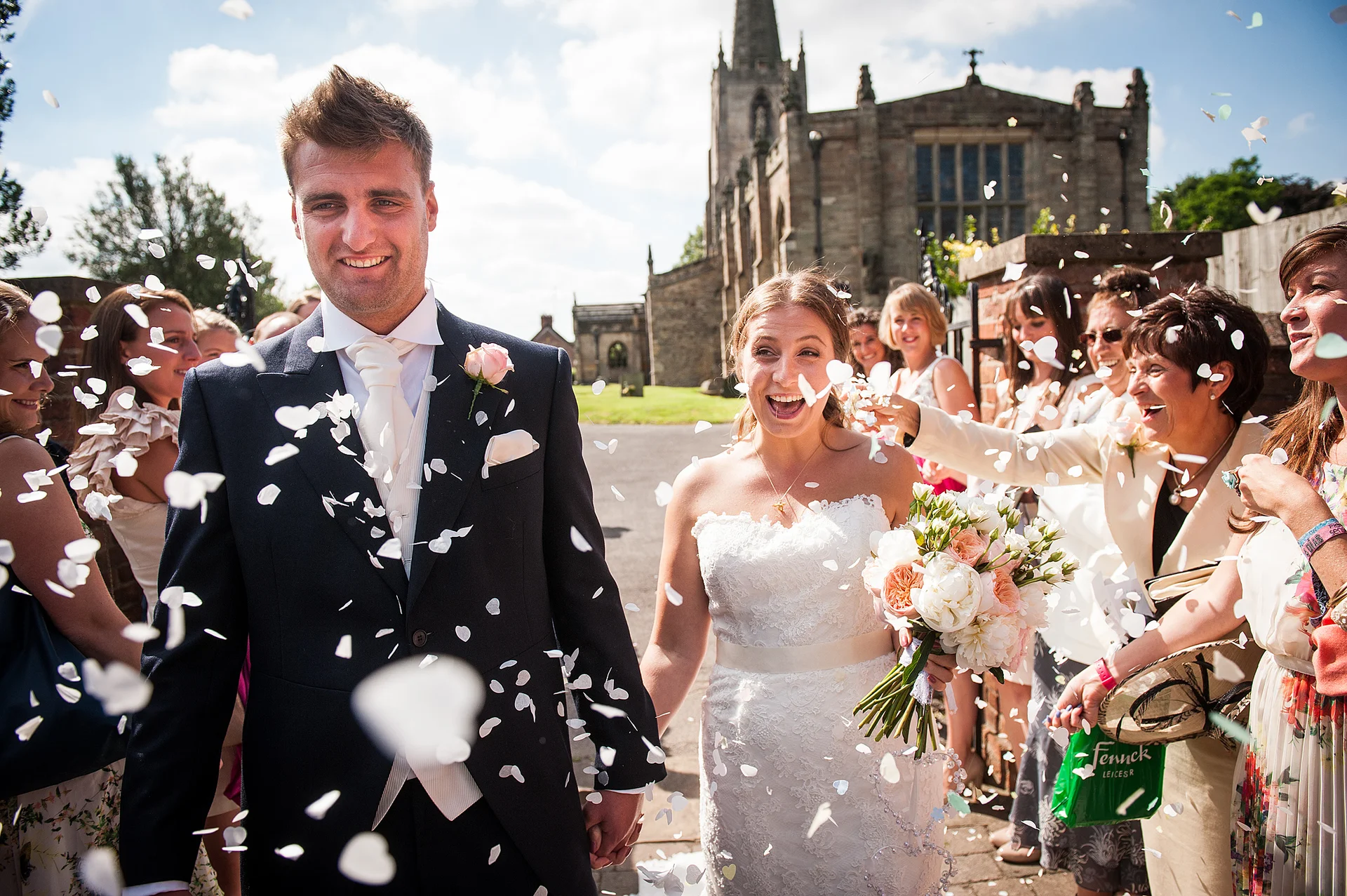 A joyful bride and groom walk hand in hand out of a traditional church while wedding guests shower them with cascading confetti petals.