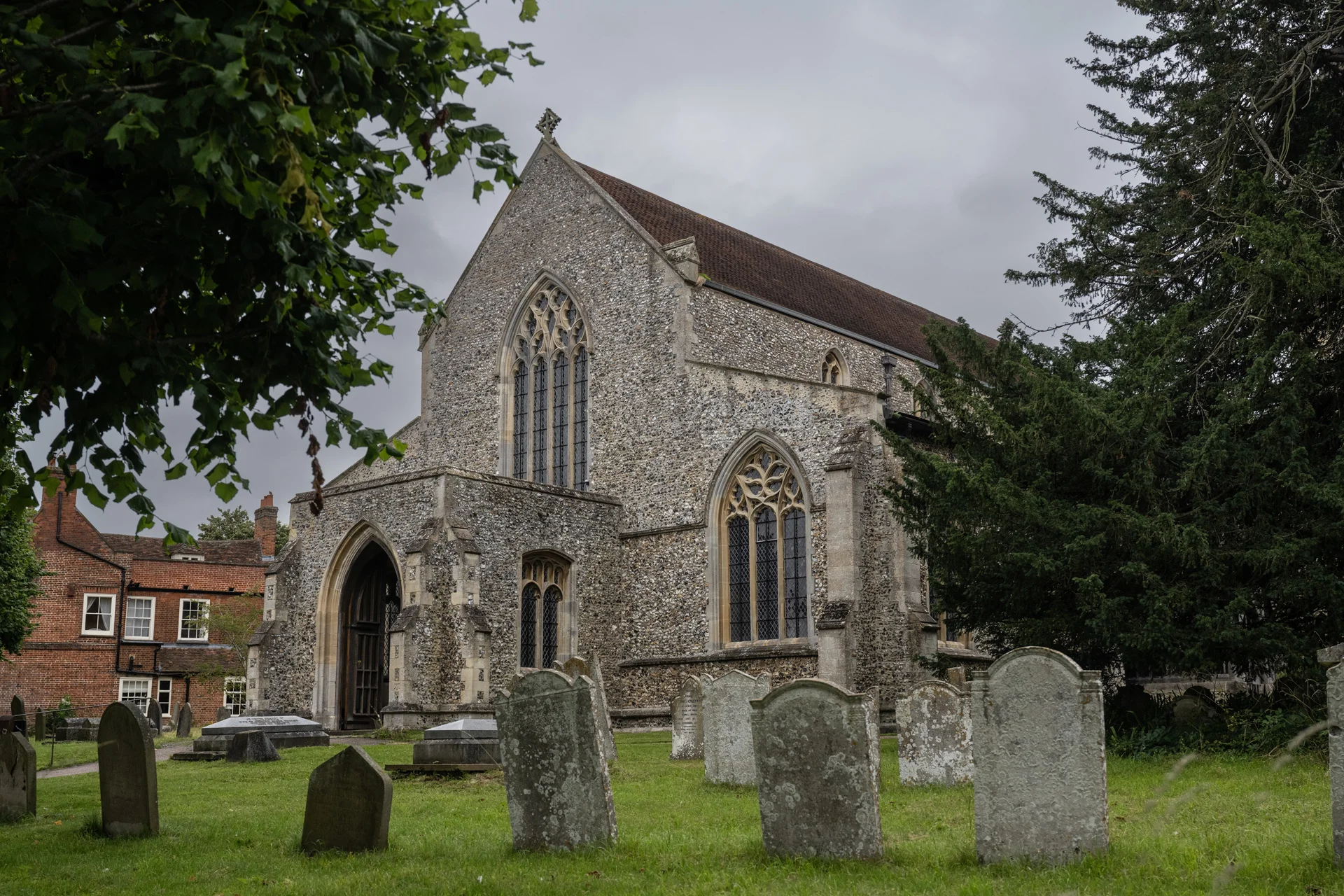 An exterior view of a historic stone church surrounded by a green churchyard with weathered headstones beneath an overcast sky.
