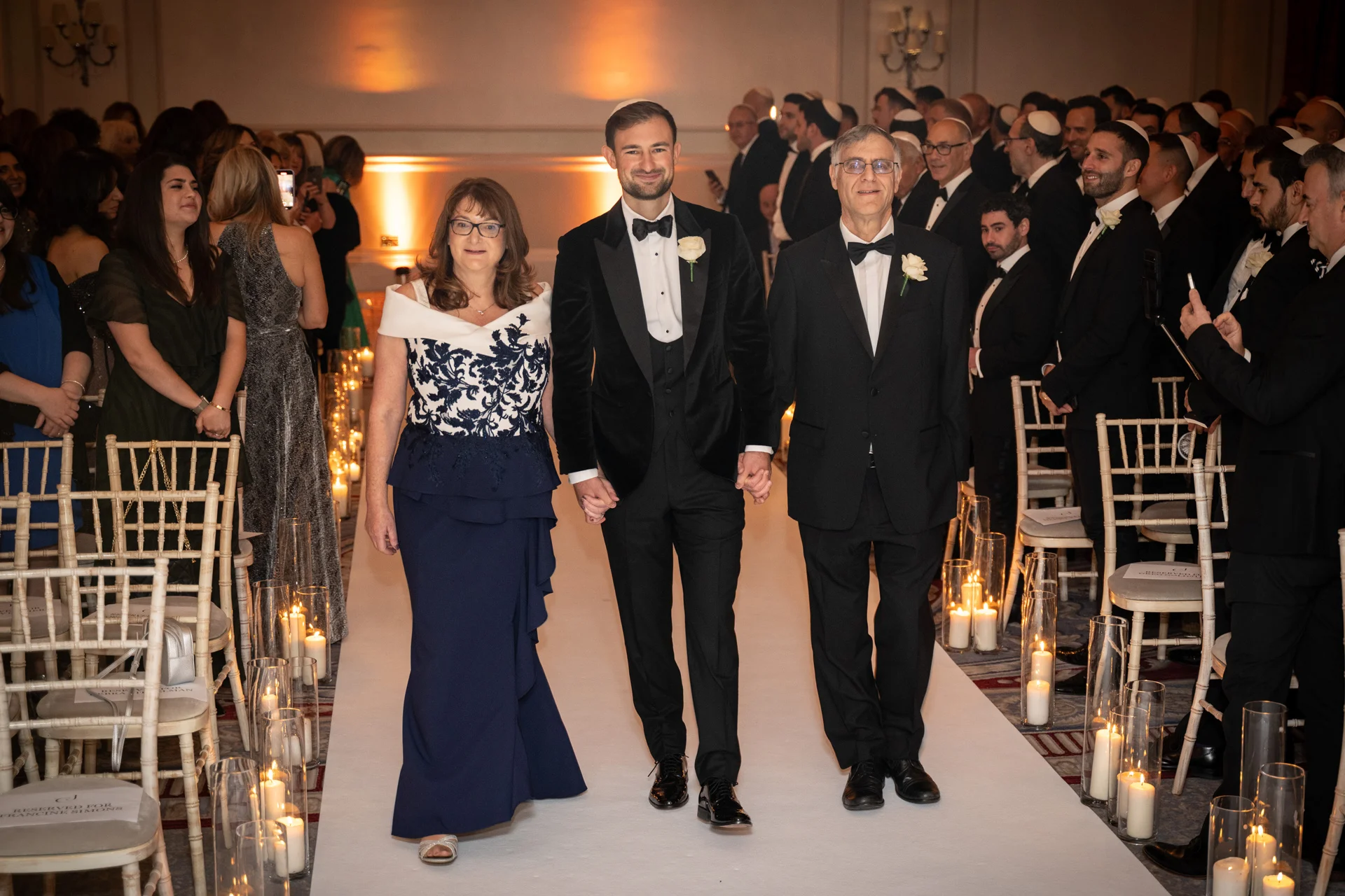 A smiling groom walks down the aisle hand-in-hand with his parents, flanked by candlelit lanterns and rows of seated guests in a formal ballroom.