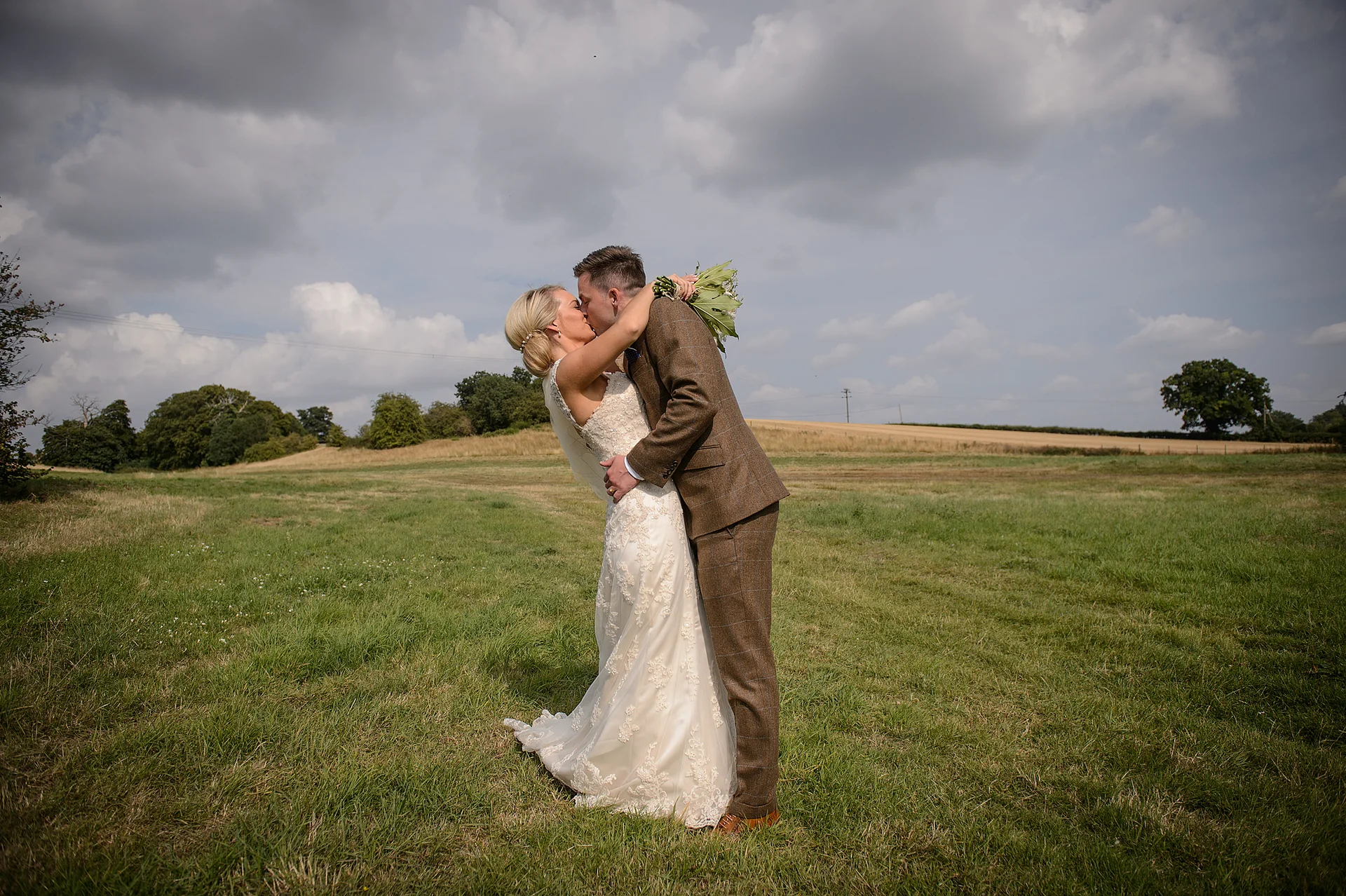 A newlywed couple shares a romantic dip kiss in the middle of a grassy field under a dramatic cloudy sky.