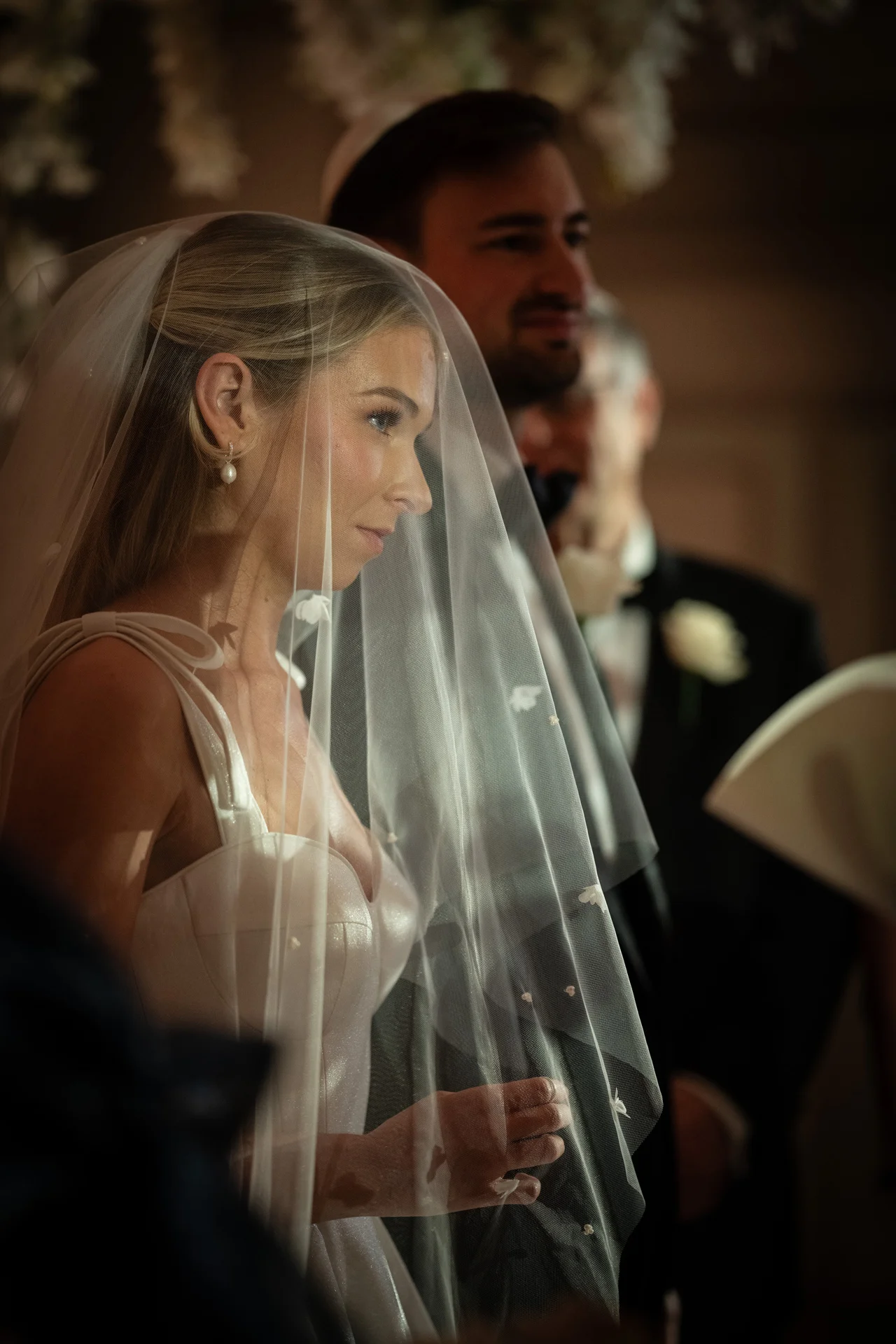 A beautiful bride stands beneath a chuppah during a Jewish wedding ceremony, wearing a structured gown and a sheer veil draped over her face.