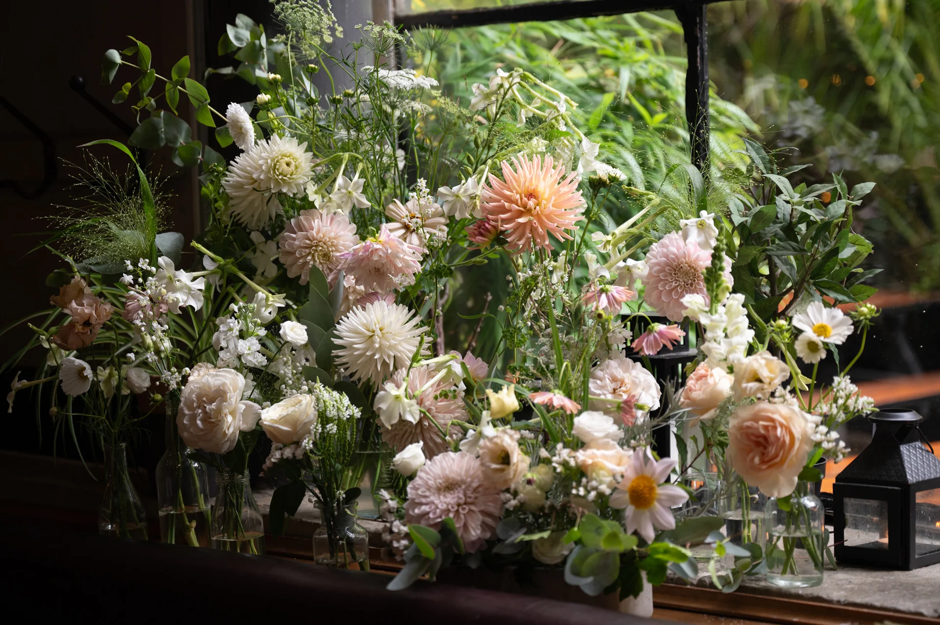 An array of delicate pastel floral arrangements featuring dahlias, garden roses, and cosmos sits in glass bud vases on a windowsill, illuminated by soft natural window light.