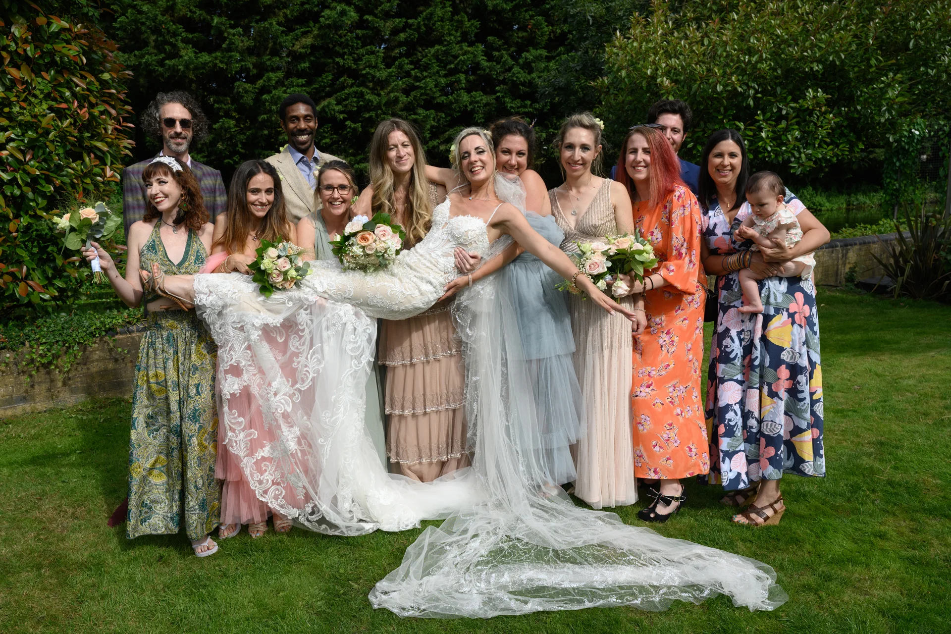 A joyful bride in a lace gown with a cathedral veil is being playfully lifted horizontally by her wedding party in a sunlit garden.