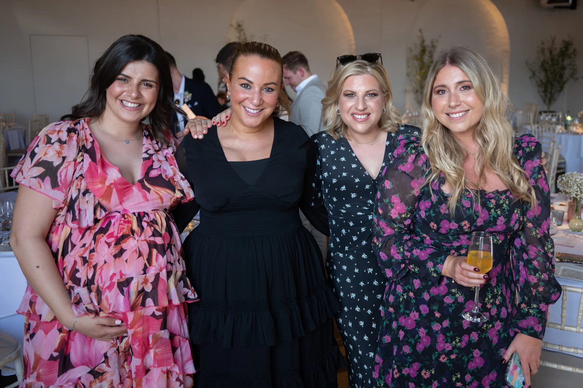 Four smiling female wedding guests pose together in a bright reception space wearing stylish floral print and black dresses.