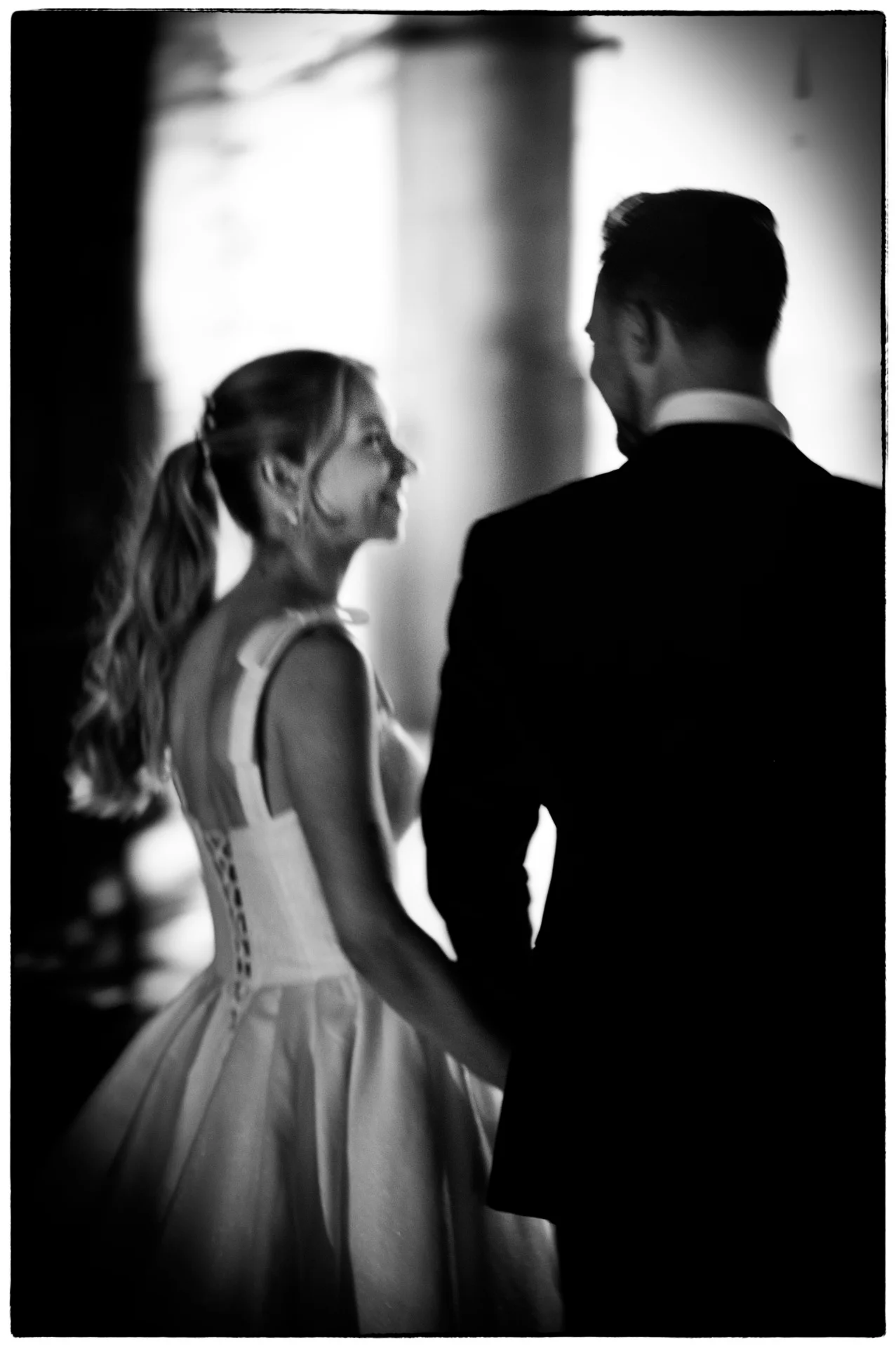 A newlywed couple walk together in a dramatic black and white photograph, with the bride looking back lovingly towards the groom.