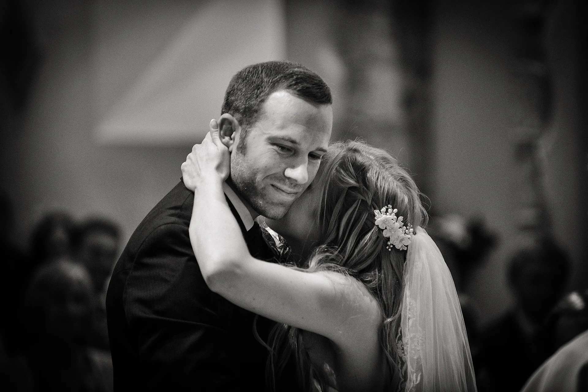 A newlywed couple shares an intimate embrace during their wedding celebration, captured in a moody black and white photograph.