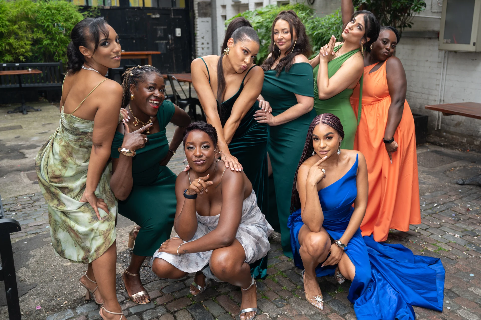 A lively group of bridesmaids and wedding guests pose together in a cobbled courtyard, wearing a vibrant mix of emerald green, blue, and orange dresses.