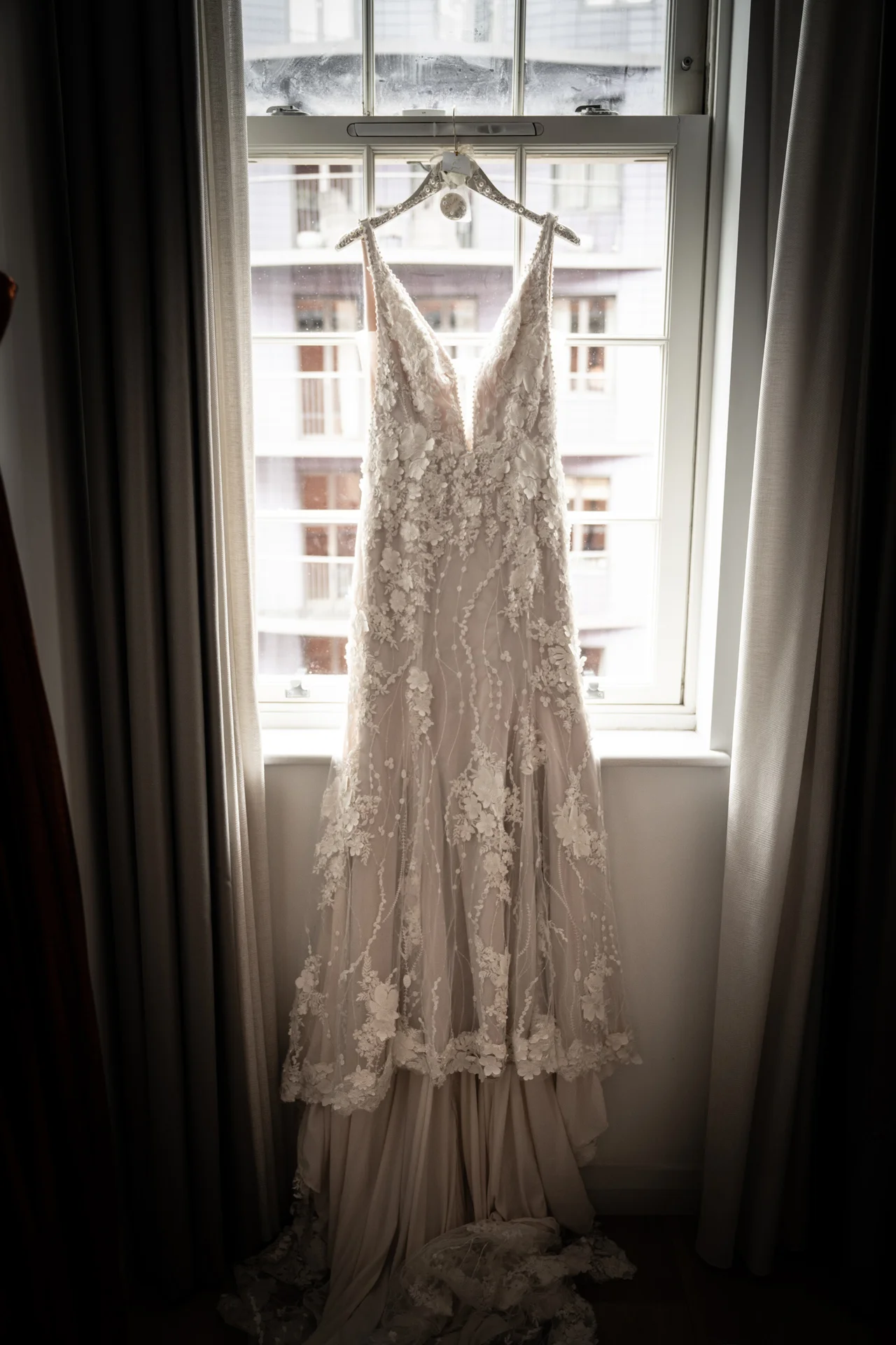 A detailed lace wedding dress hangs gracefully in front of a bright window, beautifully illuminated by natural light during bridal preparations.