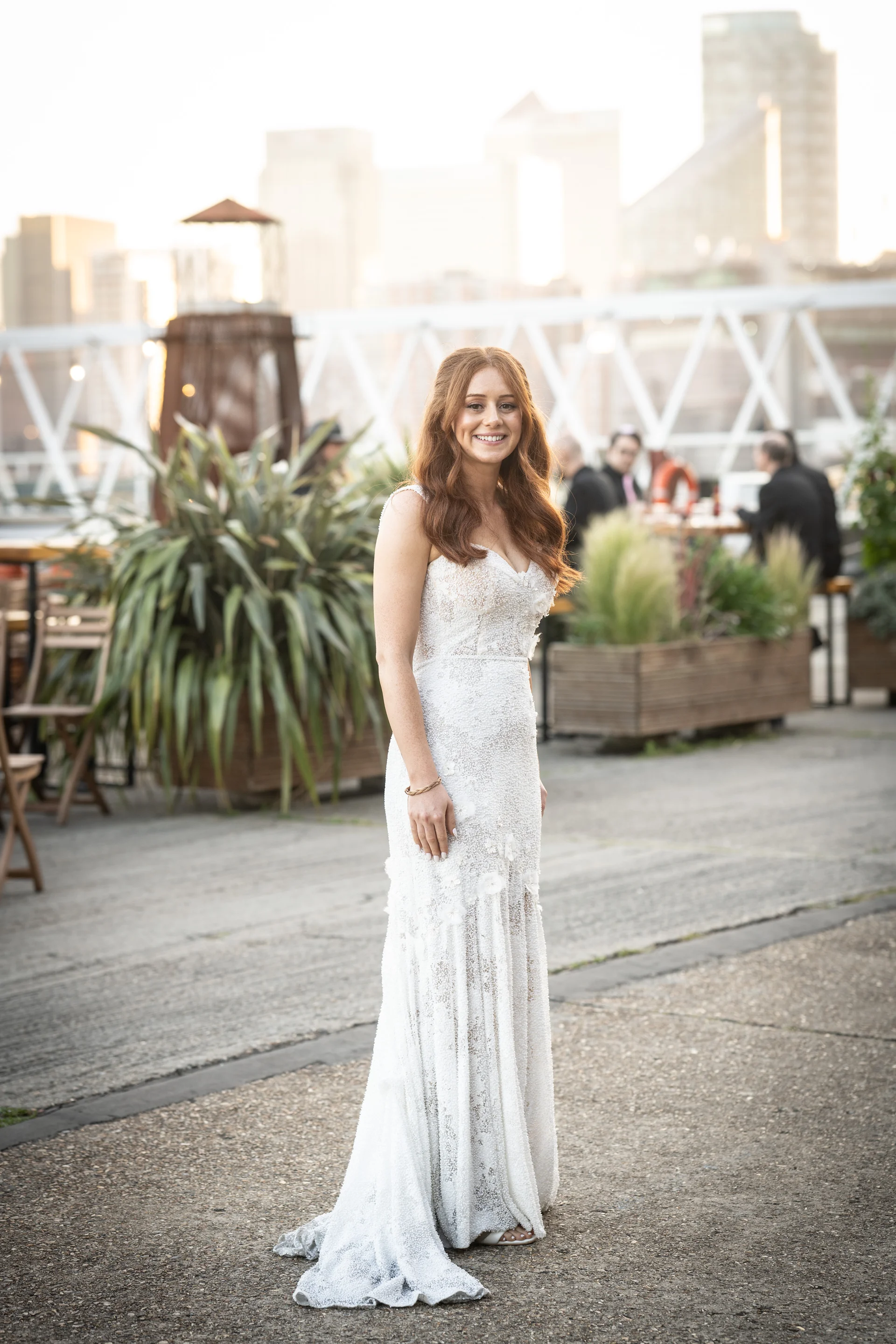 A stunning red-haired bride poses gracefully in a beaded lace sweetheart gown on a scenic rooftop terrace with a city skyline backdrop during golden hour.