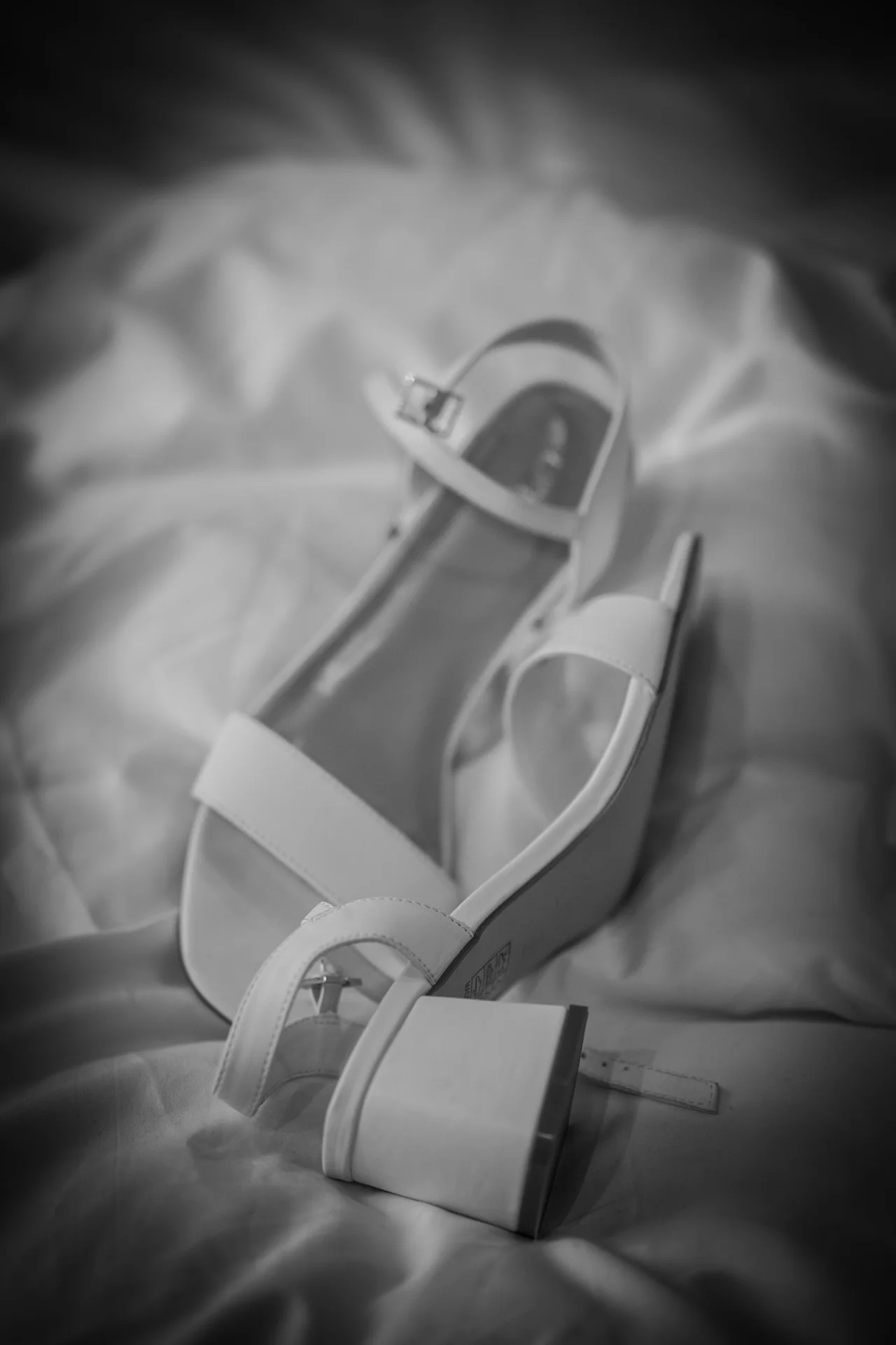 A stylish black and white detail shot captures a pair of elegant bridal block heel sandals resting softly on textured bedding during morning preparations.