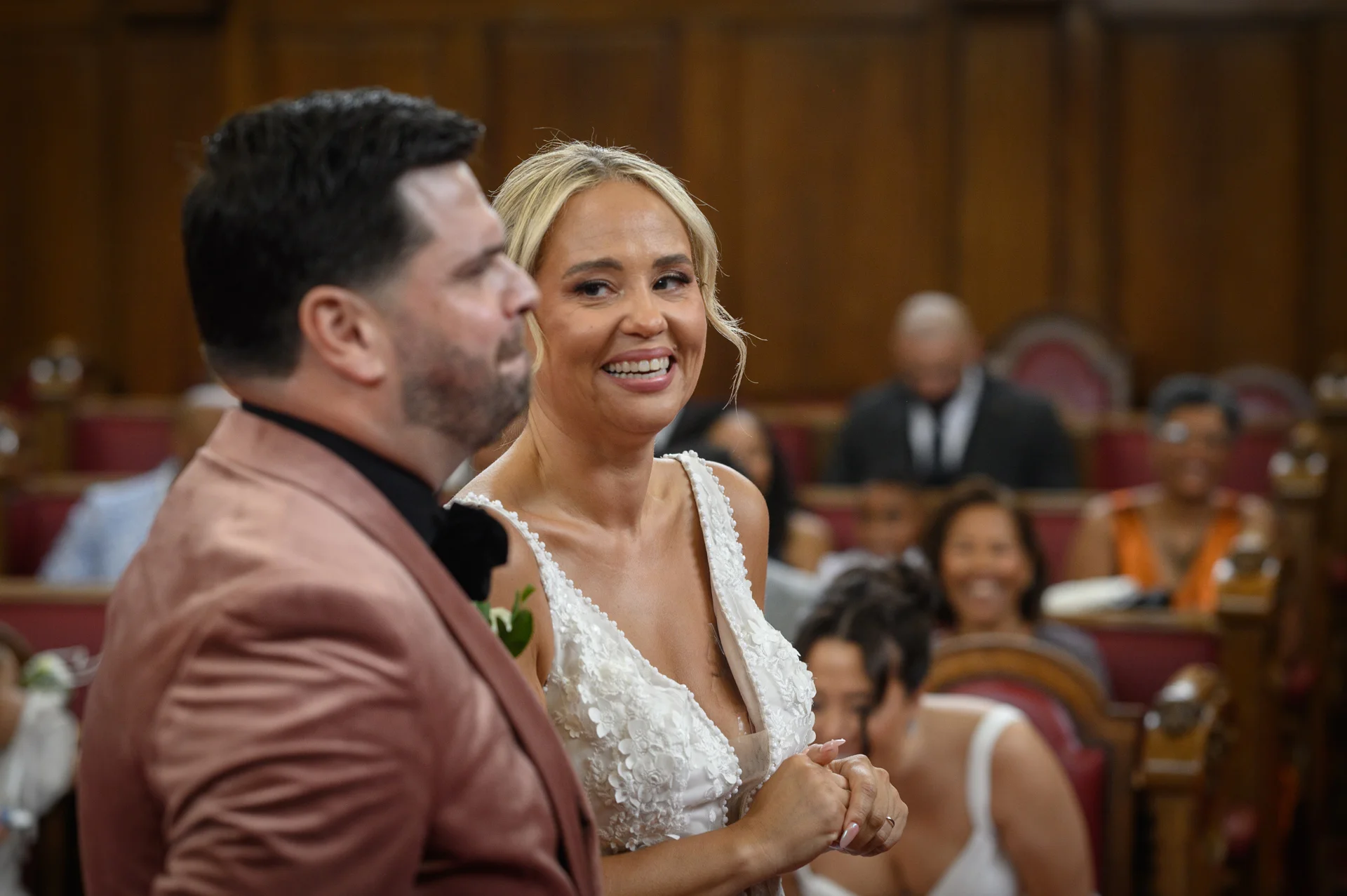 A smiling bride and groom stand together in a wood-panelled ceremony room, sharing a joyful moment during their wedding.