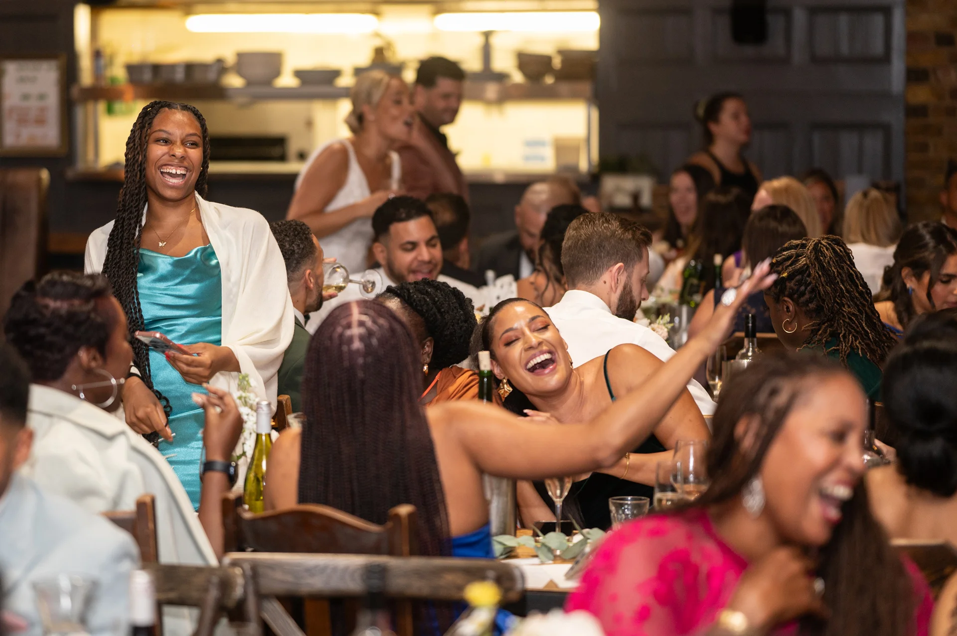 A lively group of wedding guests share a joyful moment of laughter and celebration during the wedding breakfast inside a restaurant reception.